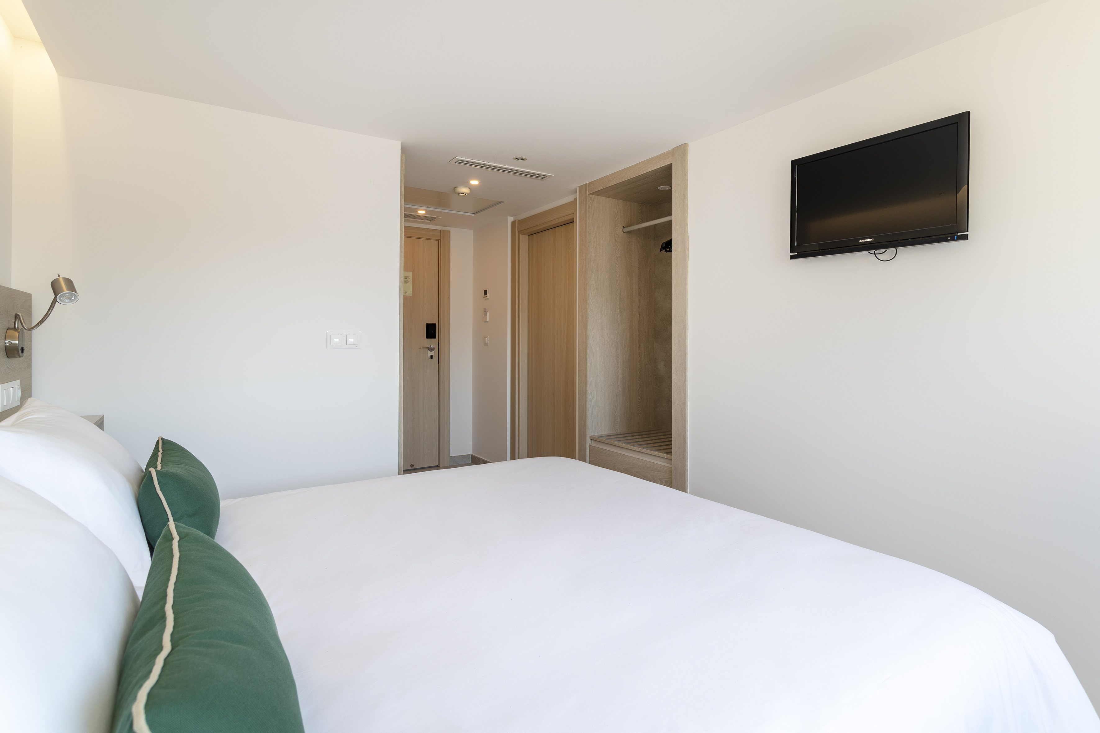 Modern hotel room with white bed, green pillows, wall TV, and closet.