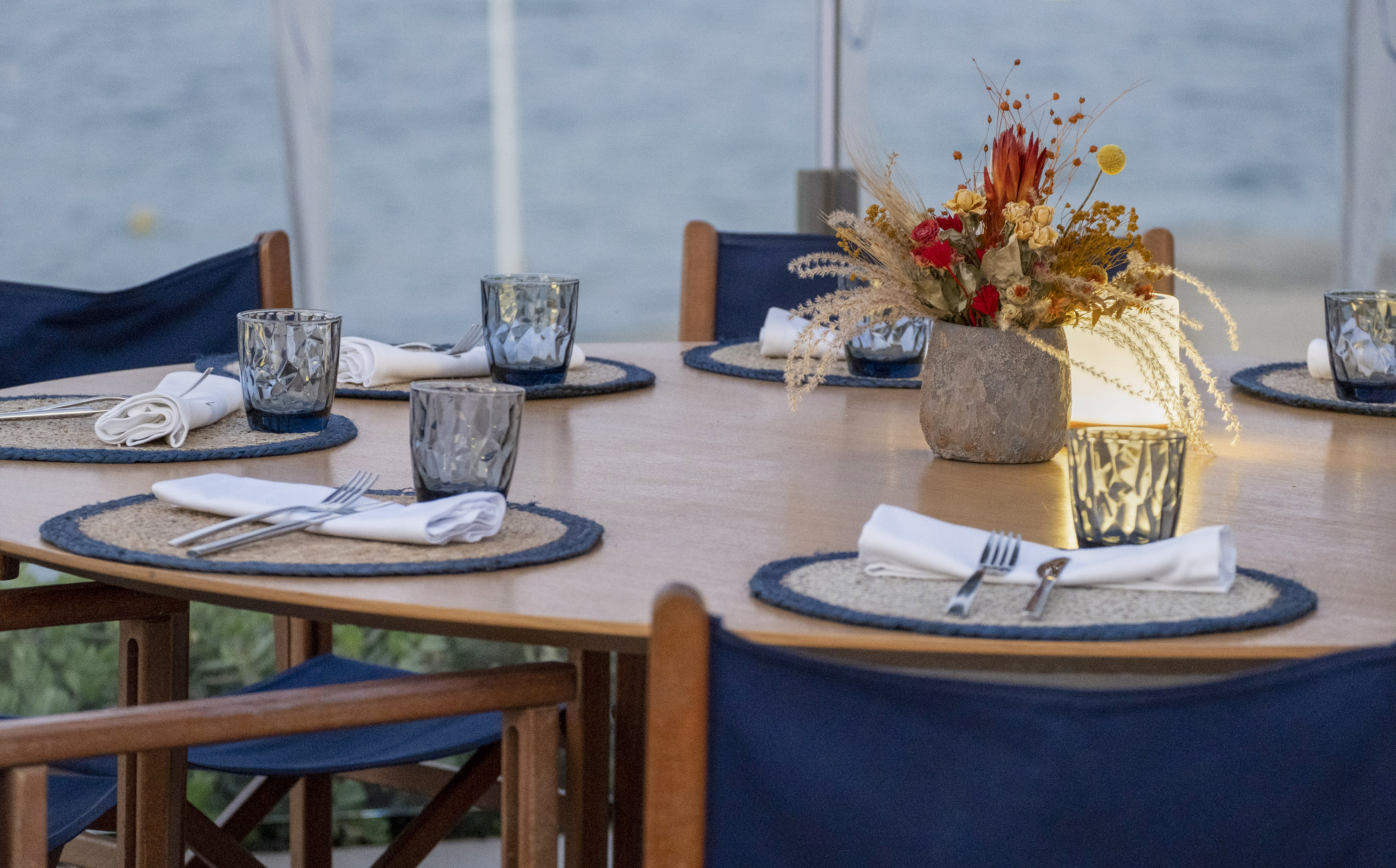 Outdoor table setting with blue details, floral centerpiece, and sea view.