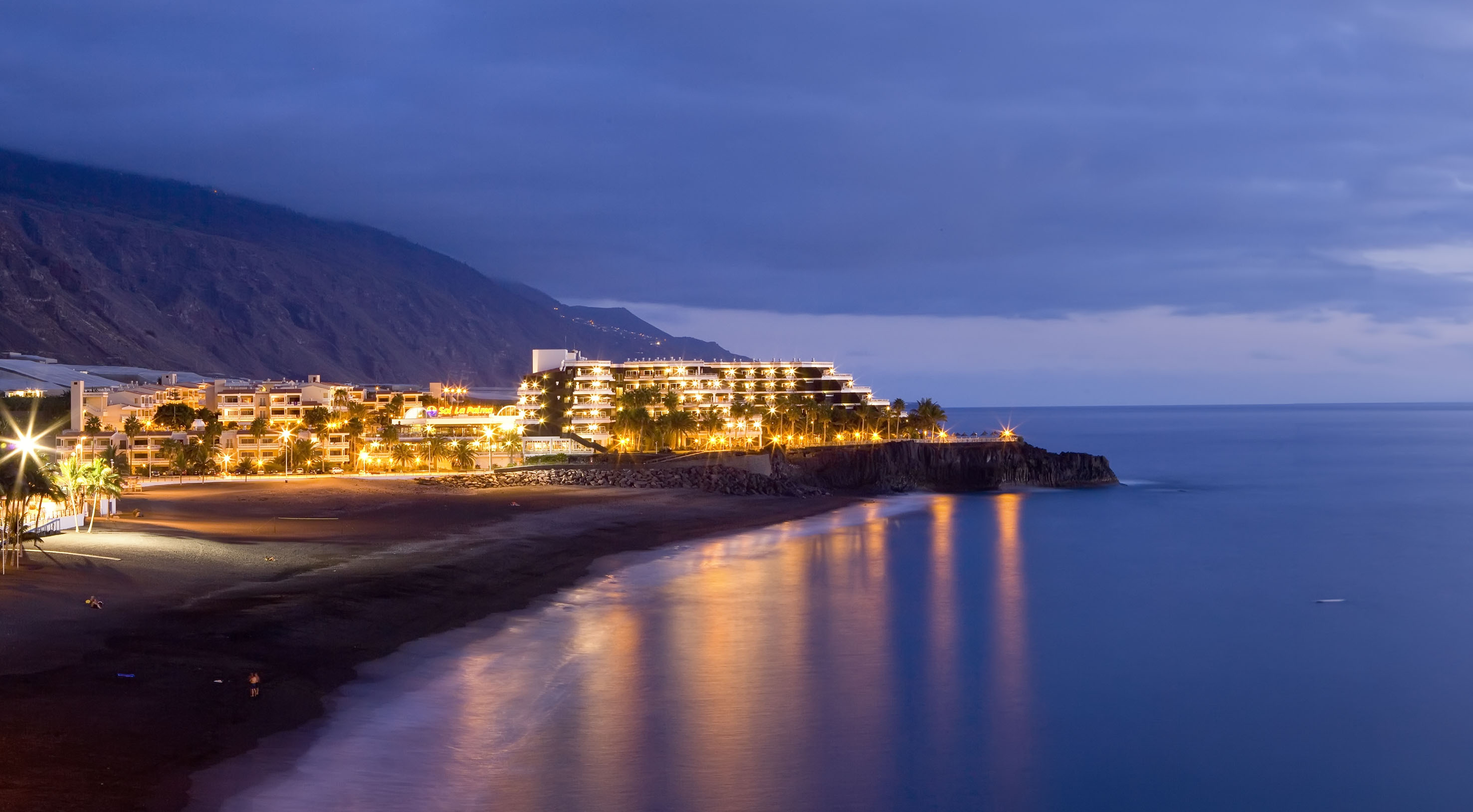 Illuminated beachfront hotel at dusk with ocean reflections and distant mountains.