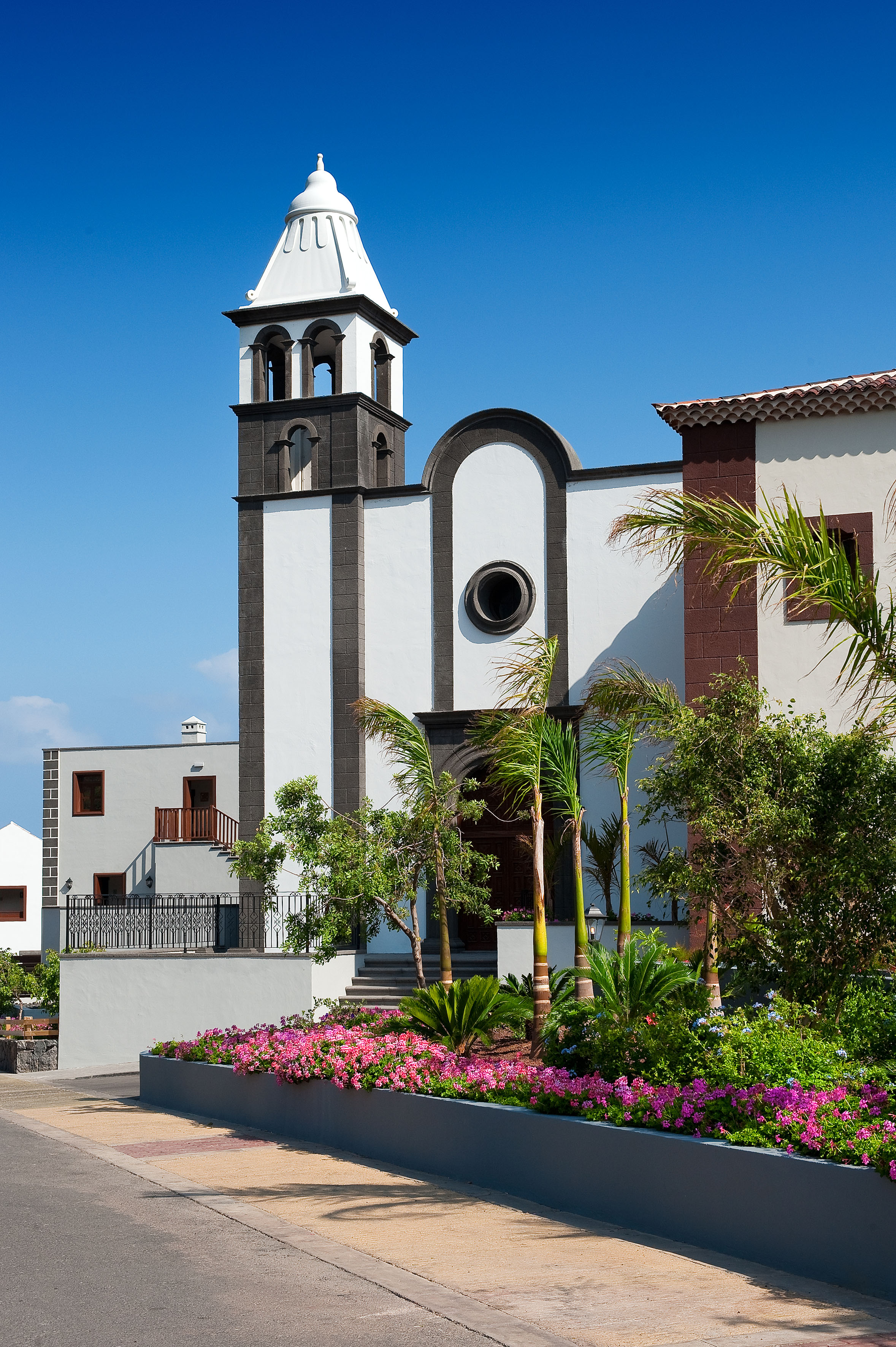 White church with black stone bell tower, palm trees, and pink flowers against blue sky.