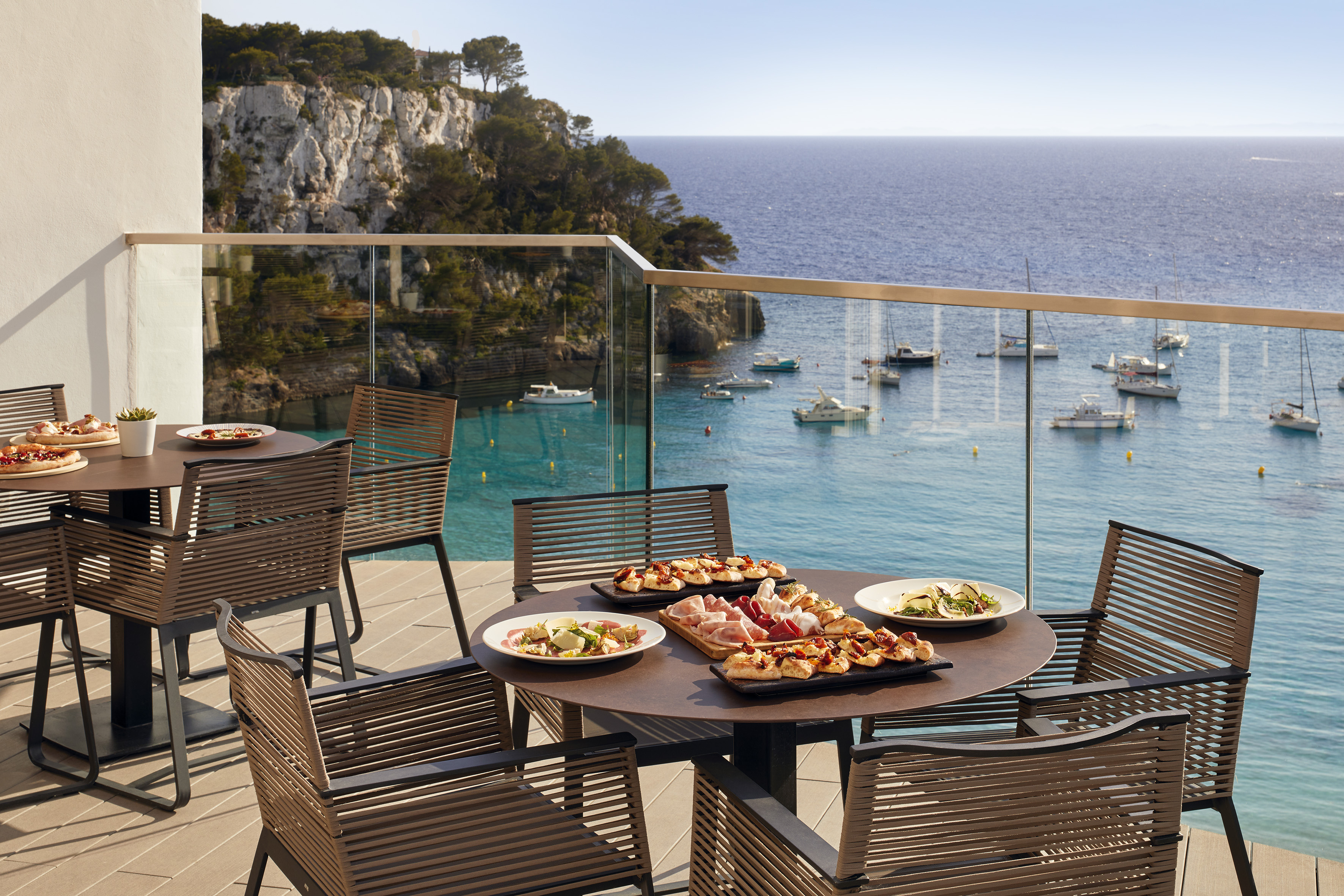 Balcony dining overlooking blue sea with boats, pizzas, and charcuterie.