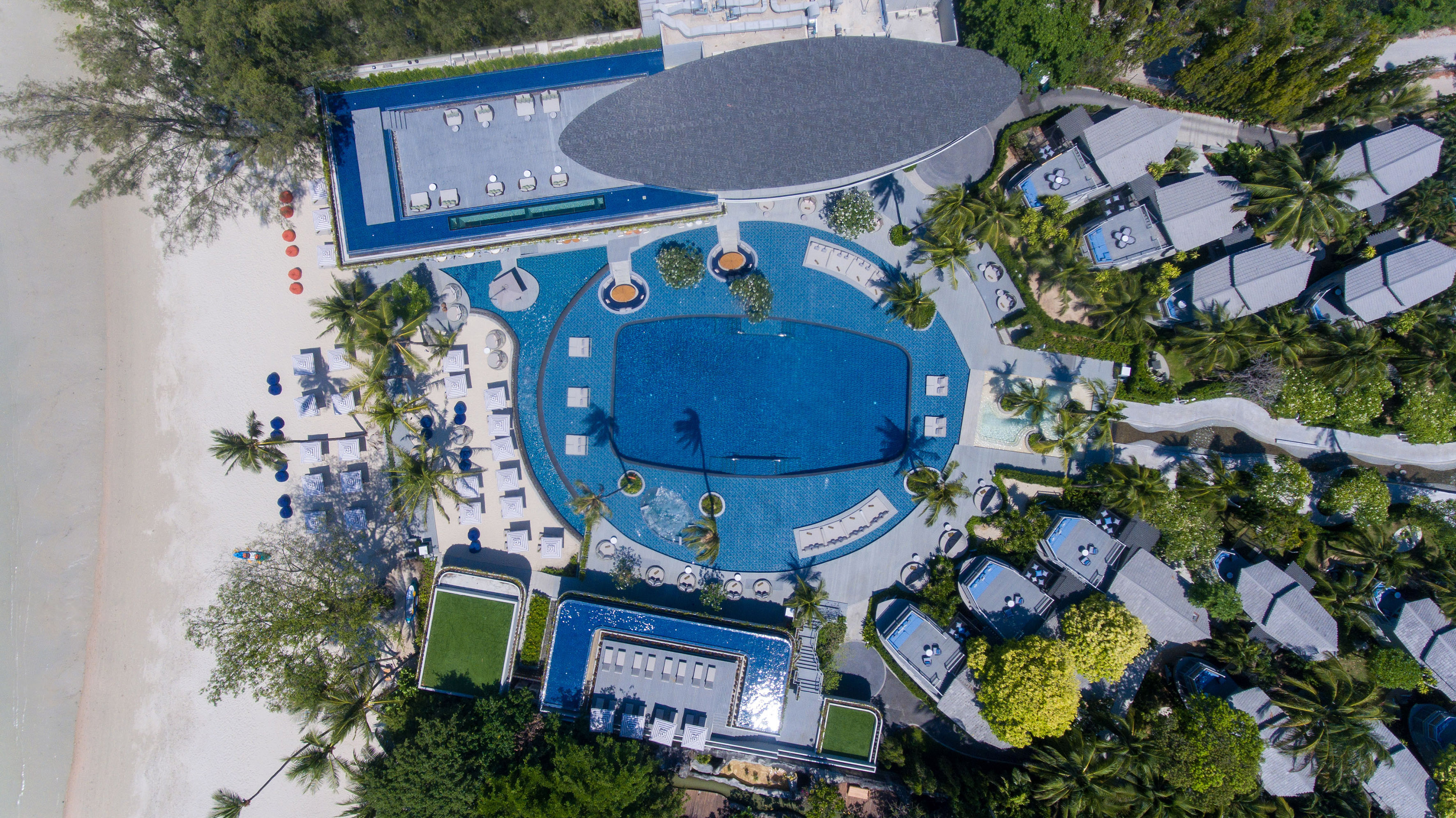 Tropical beach resort aerial: large pools, villas, sandy beach, palm trees.