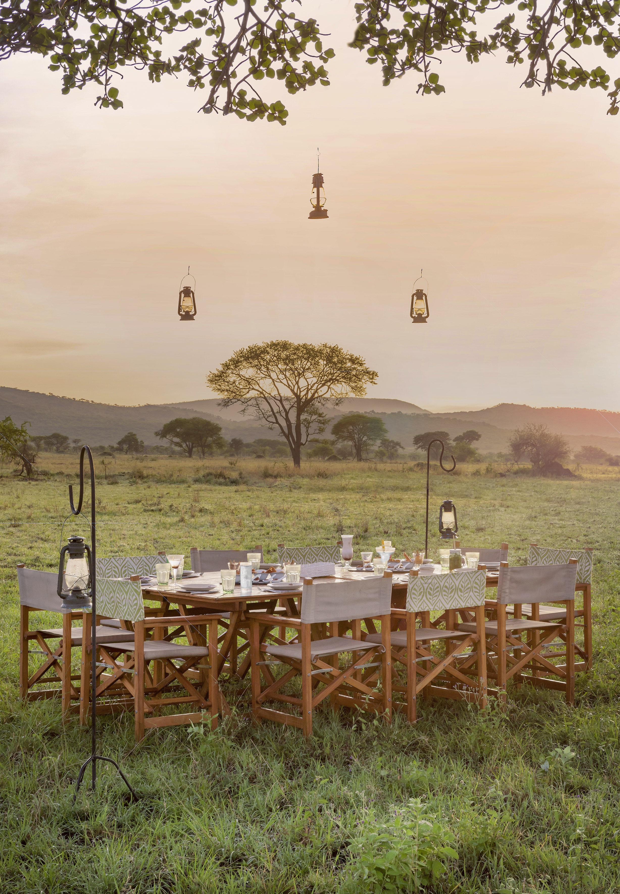 Safari outdoor dining table with lanterns in a savanna landscape at sunset.