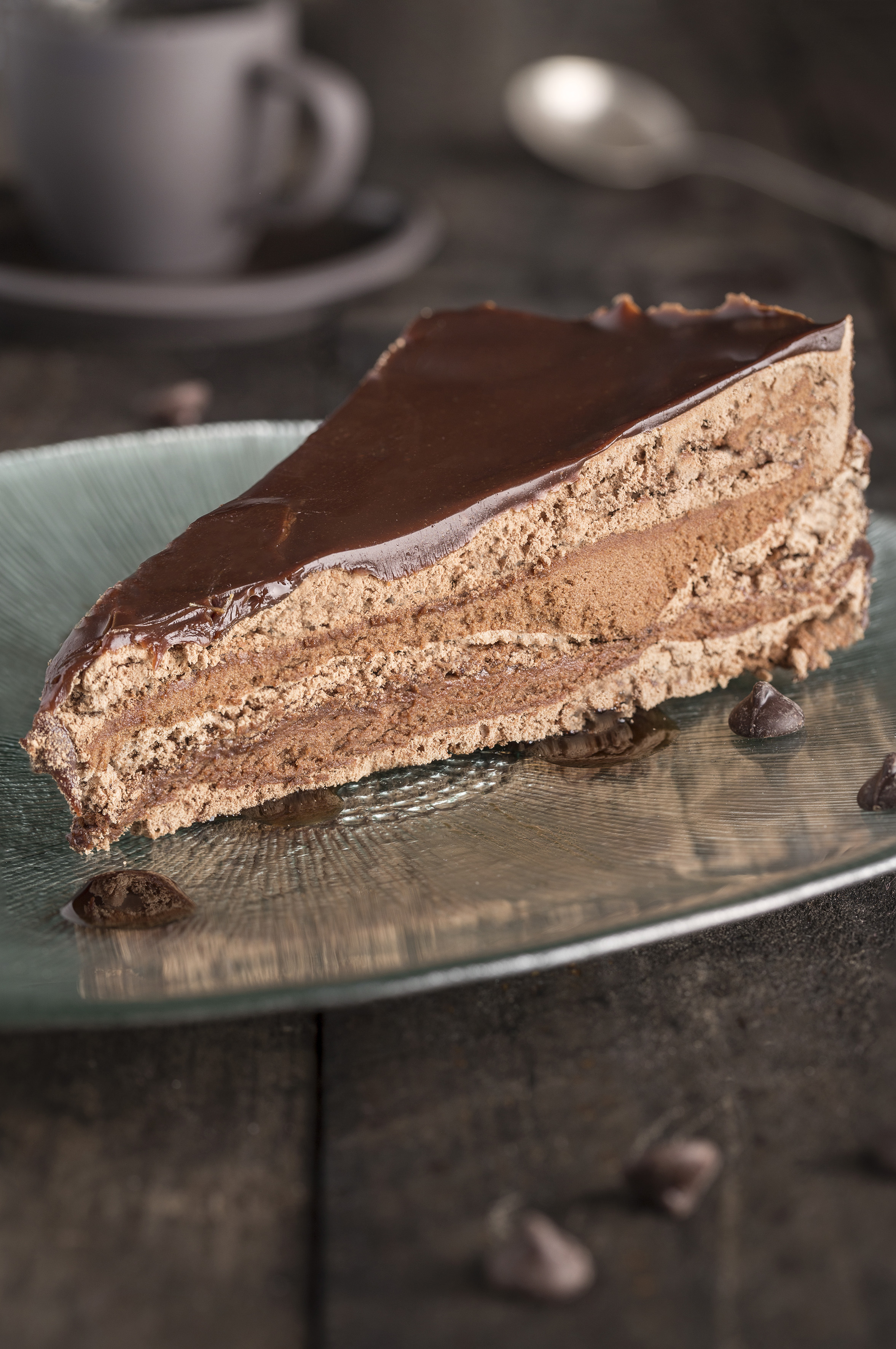 Layered chocolate cake slice with ganache on a plate, coffee in background.