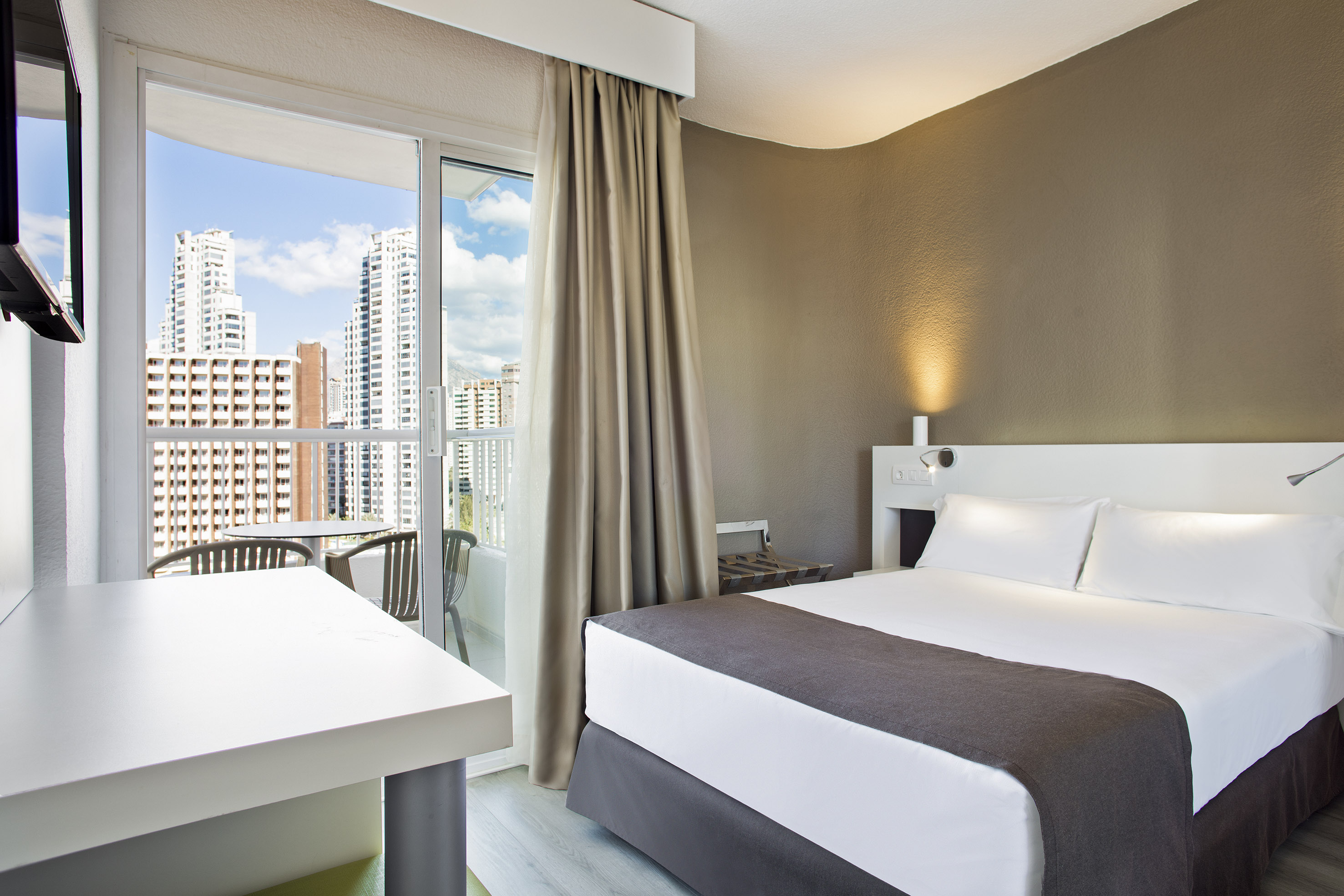 Modern hotel room with balcony city view, comfortable bed, and desk.