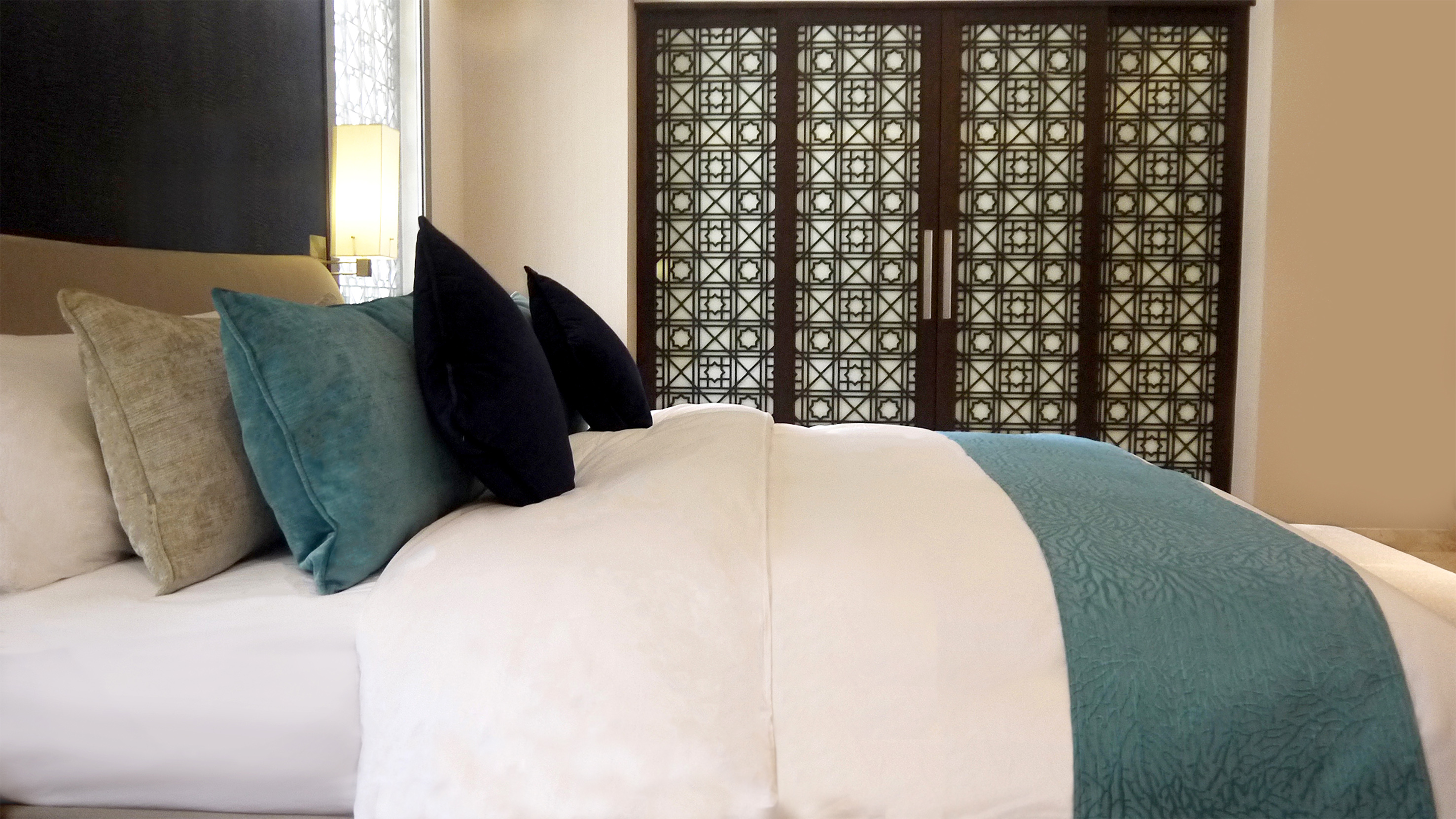 Elegant bedroom with teal and navy pillows, white bedding, geometric wooden screens.