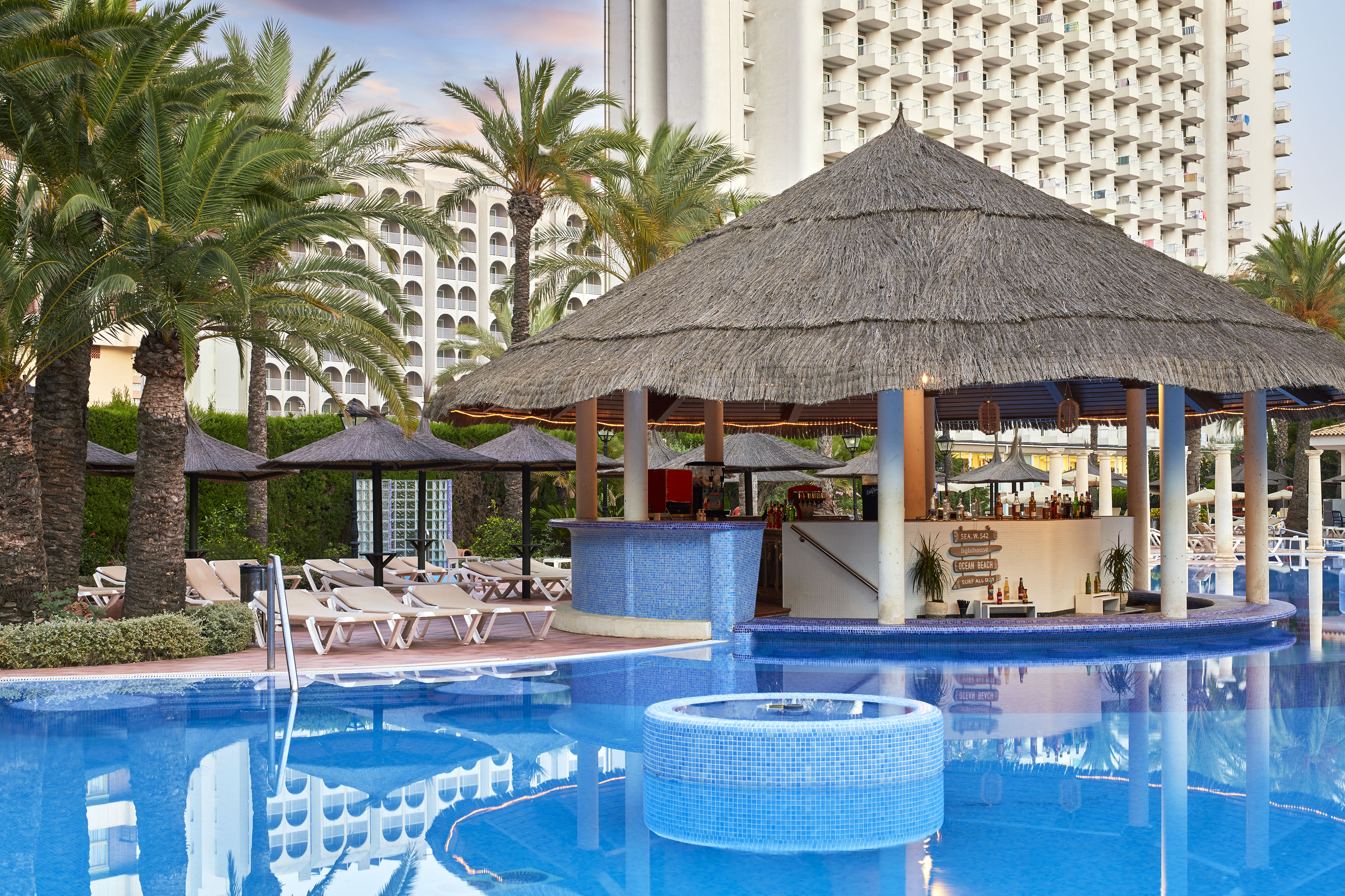 Tropical resort pool with thatched swim-up bar, lounge chairs, palm trees.