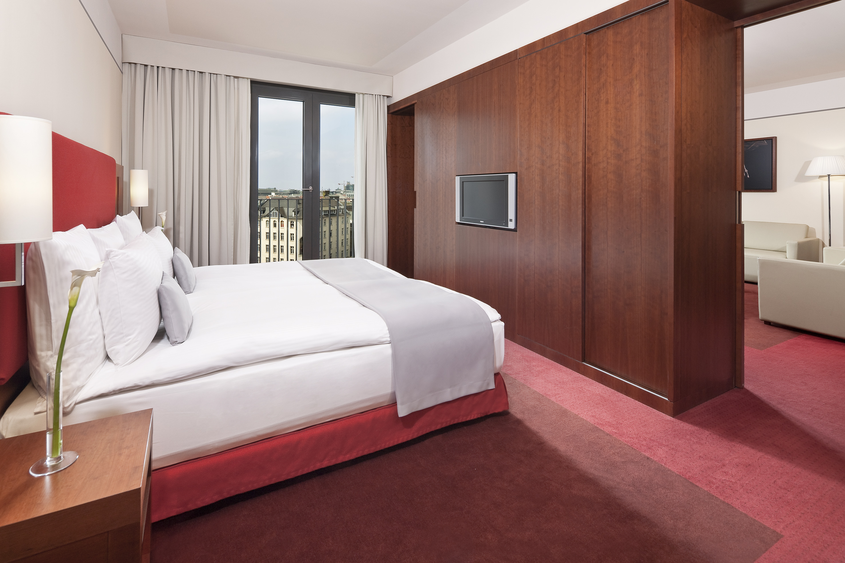 Elegant hotel bedroom with red bed, wooden wardrobe, and city view balcony.