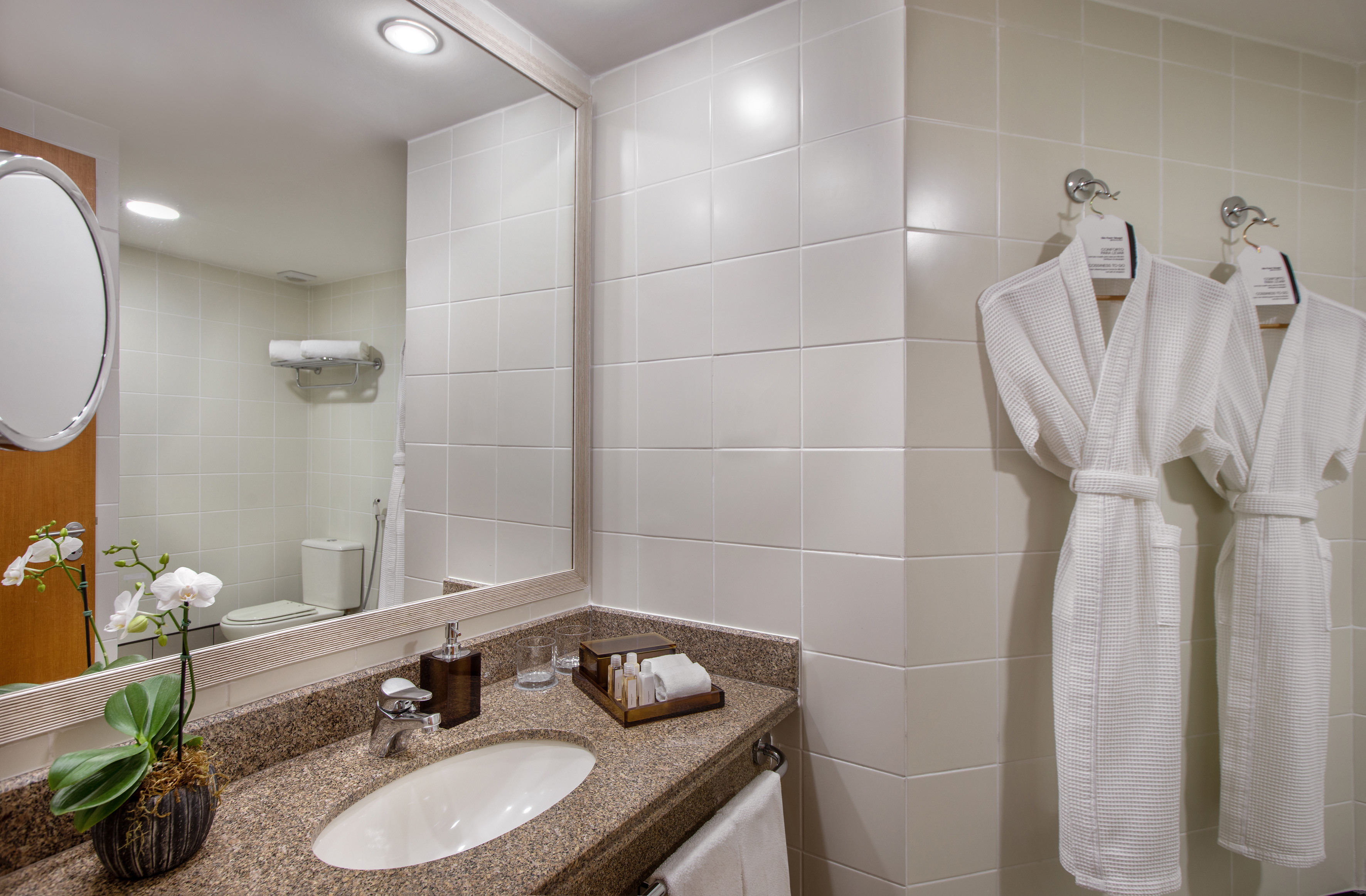 Modern hotel bathroom with granite vanity, mirror, white tiles, and two bathrobes.