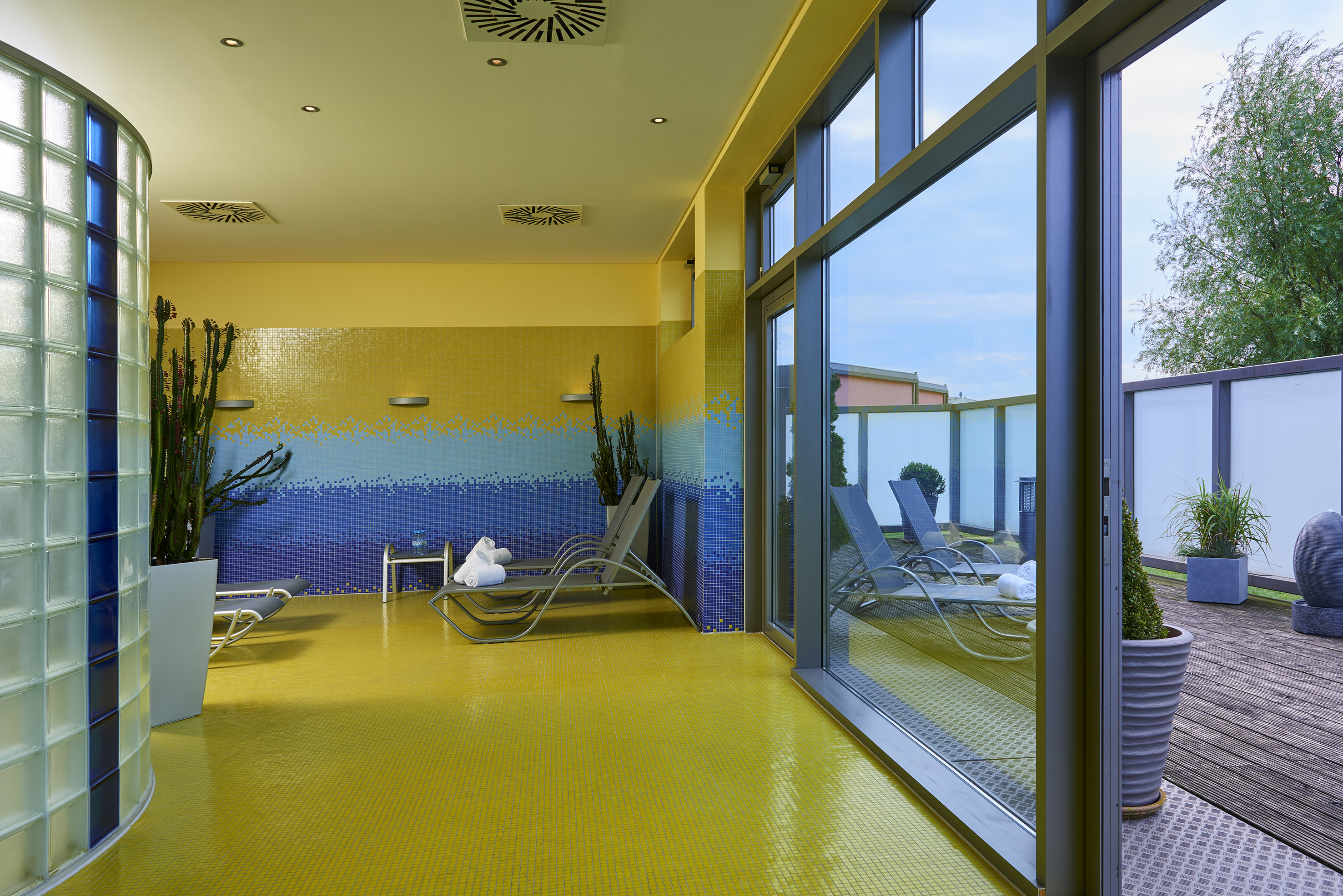 Spa lounge with yellow/blue mosaic walls, sun loungers, leading to outdoor terrace.