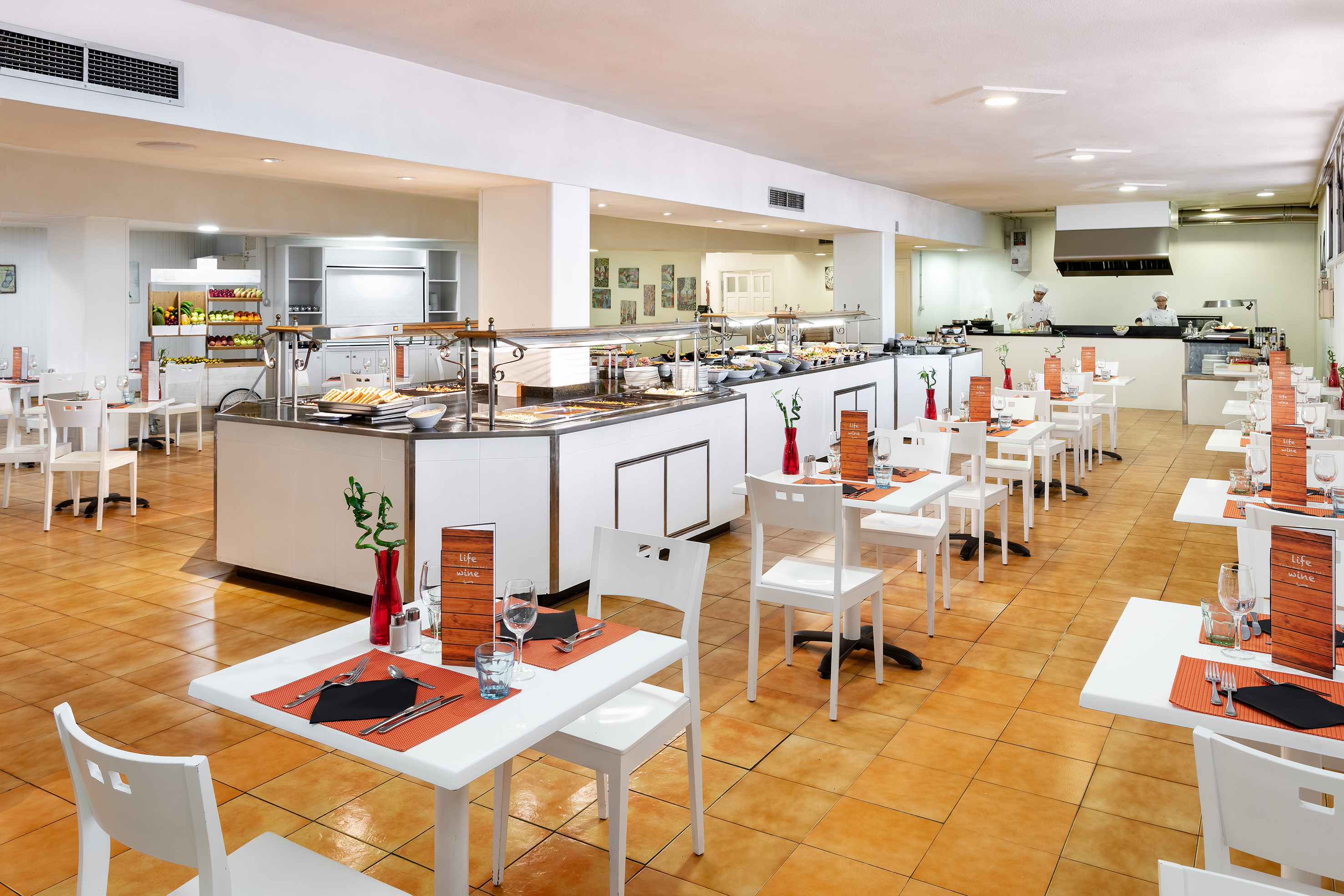 Bright hotel buffet restaurant with food stations, white tables, and chefs.