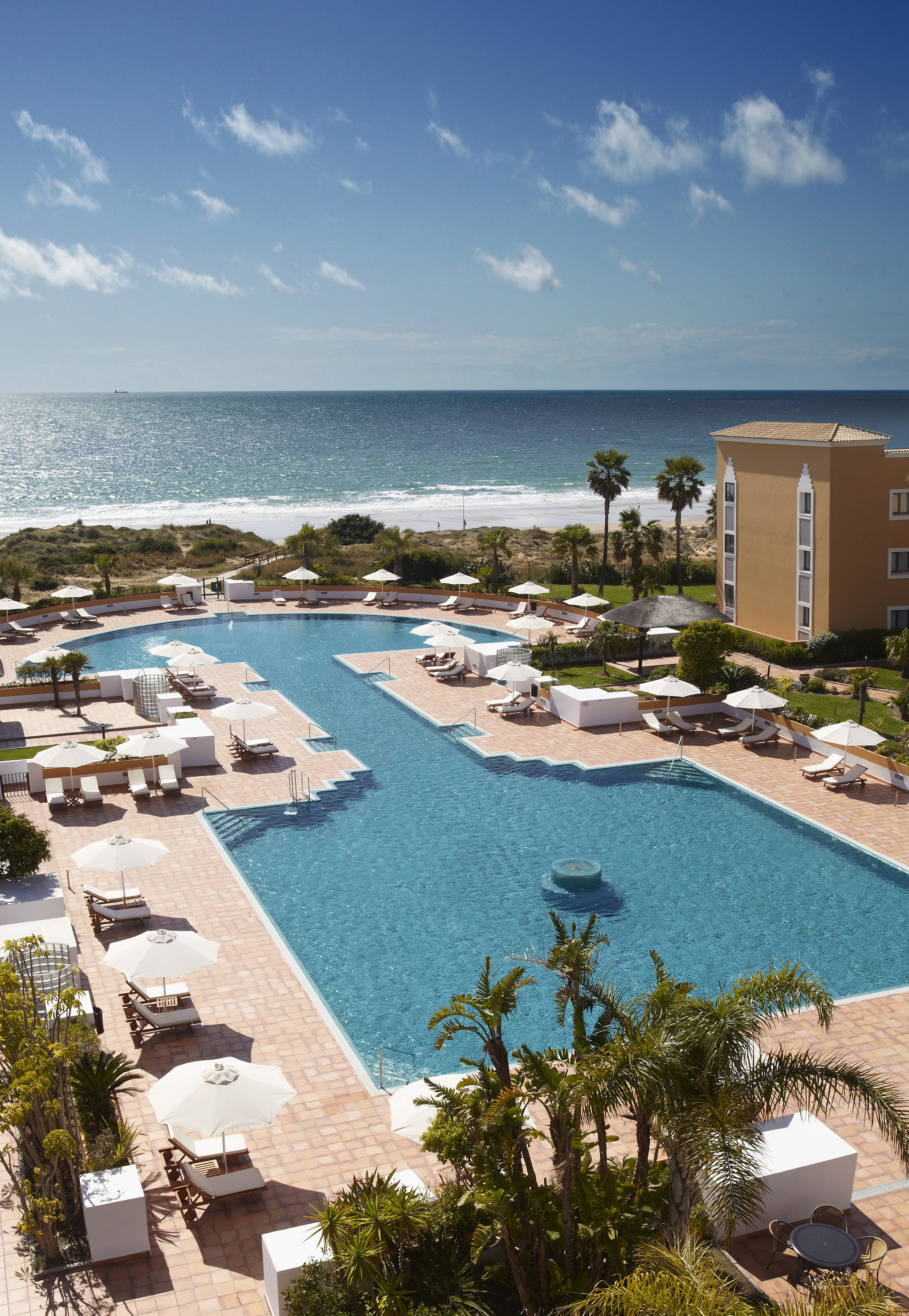 Hotel resort with large blue pool, sandy beach, and ocean view.