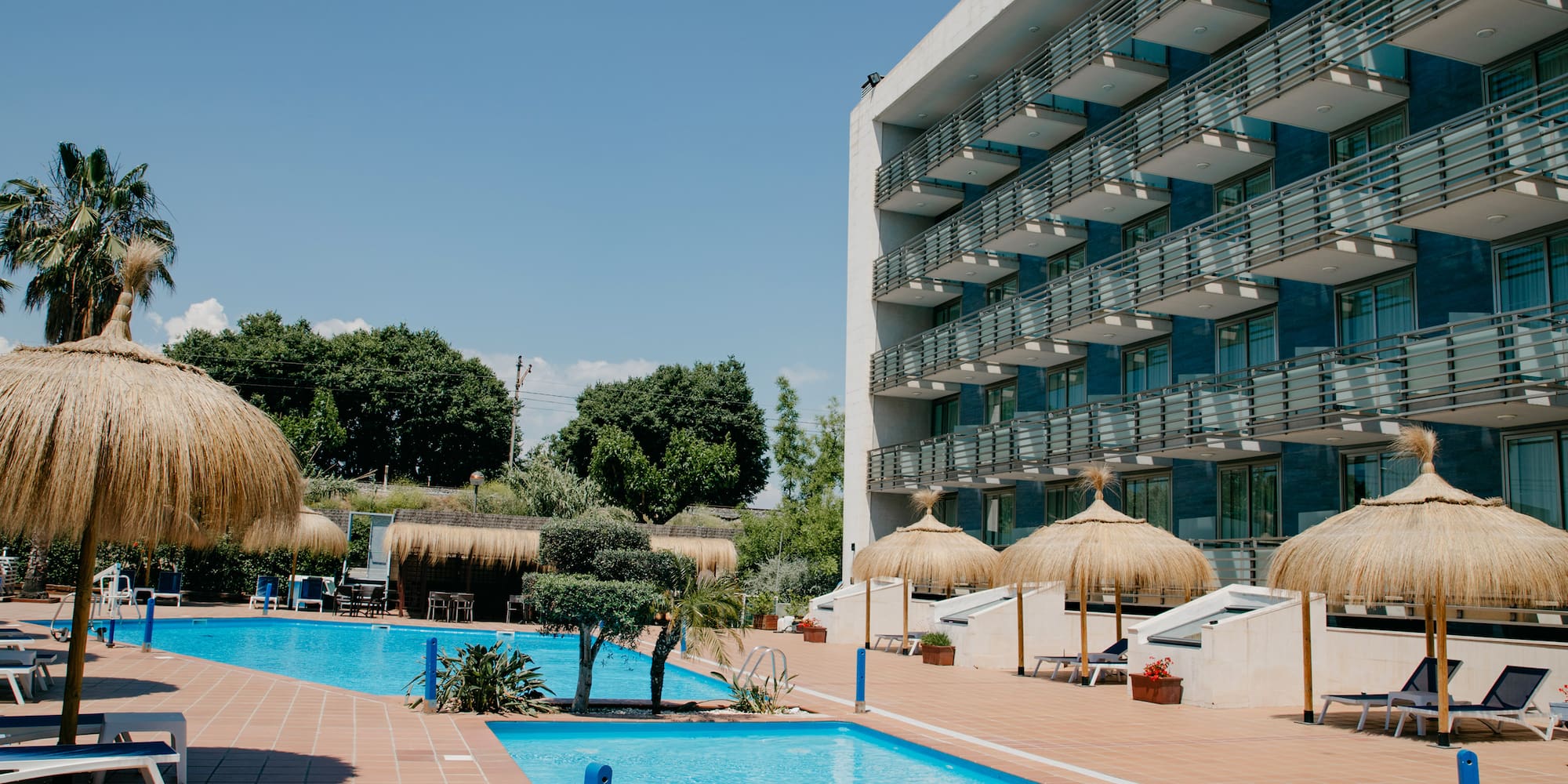 a pool with straw umbrellas and a building with trees and blue sky