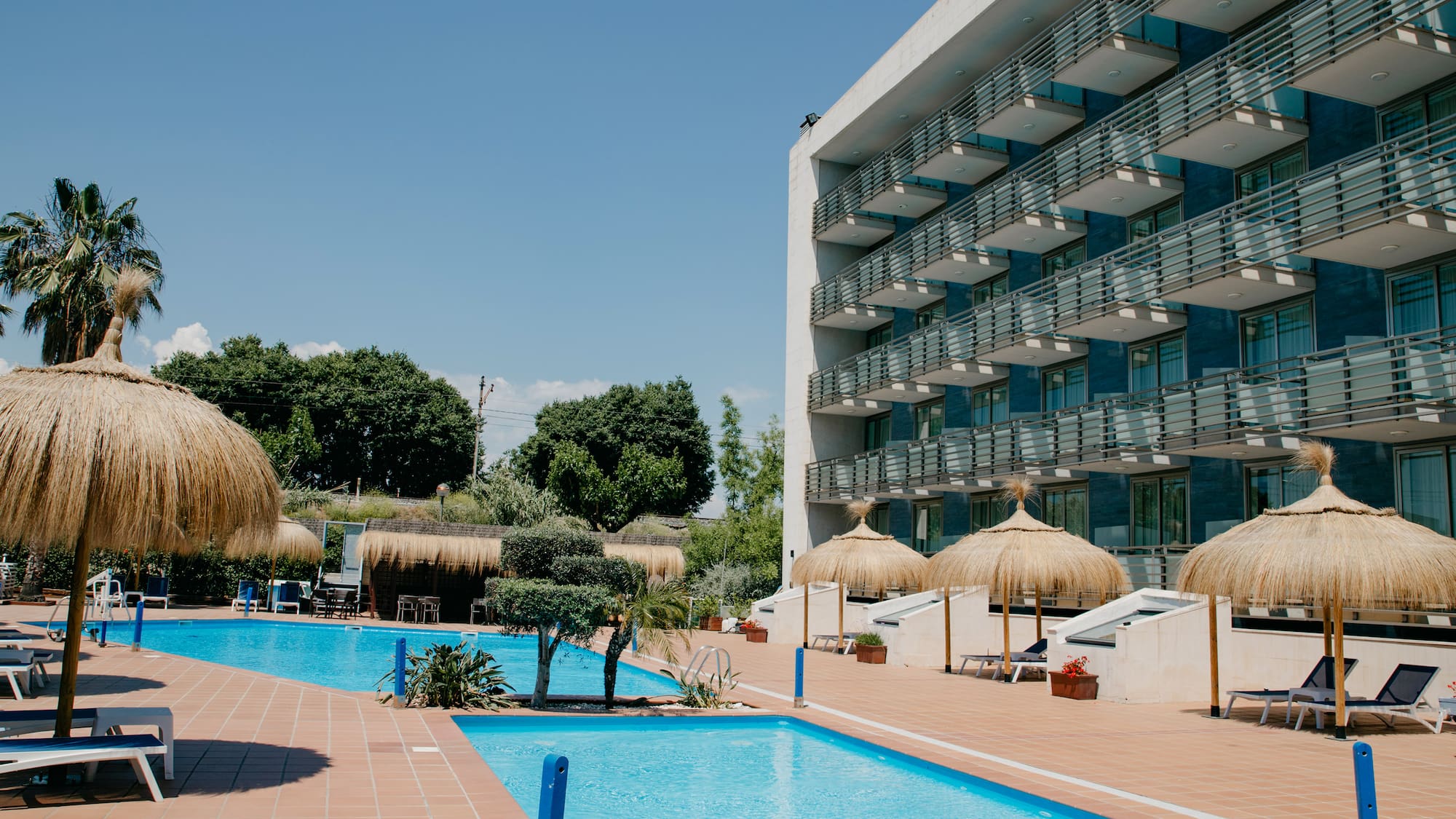 a pool with straw umbrellas and a building with trees and blue sky