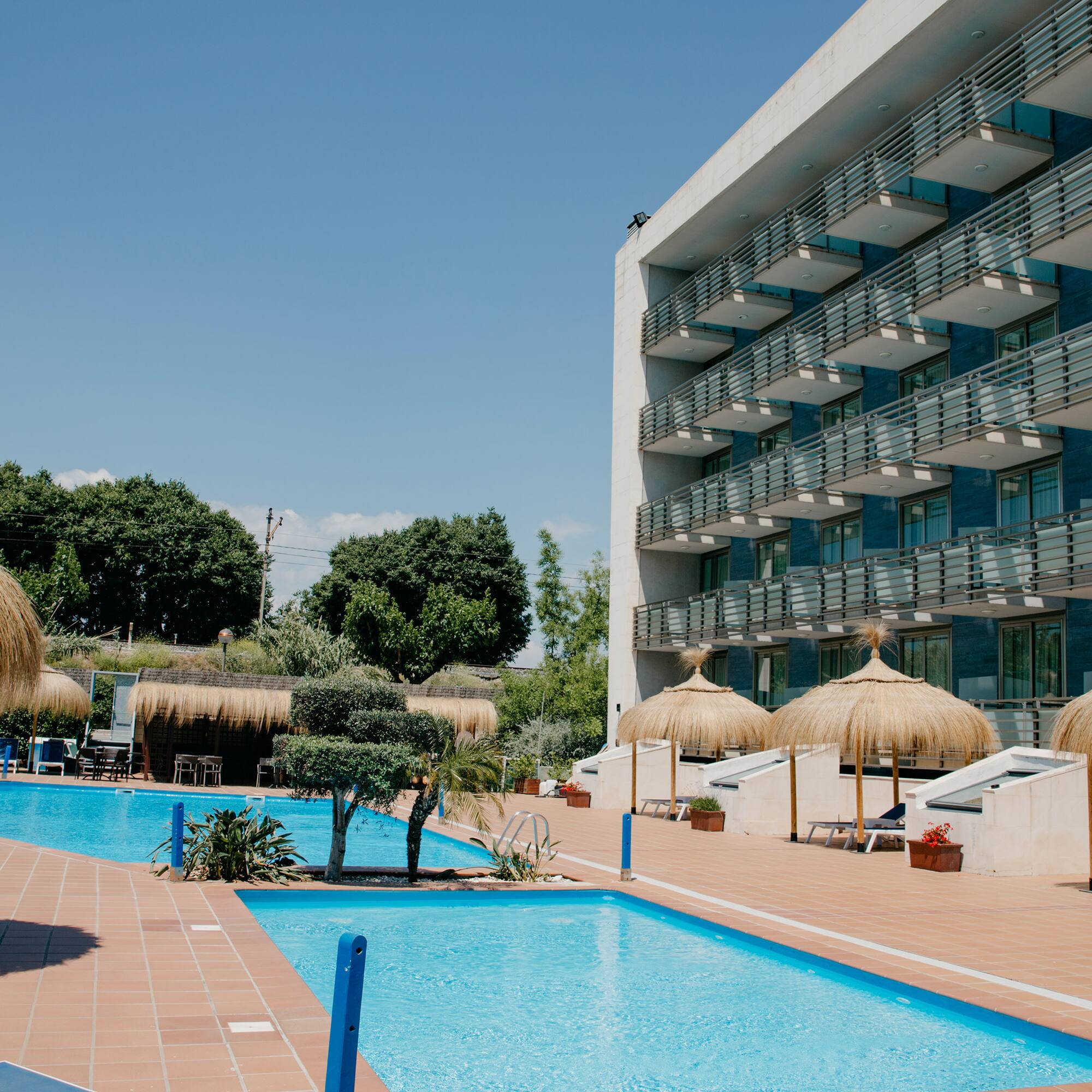 a pool with straw umbrellas and a building with trees and blue sky