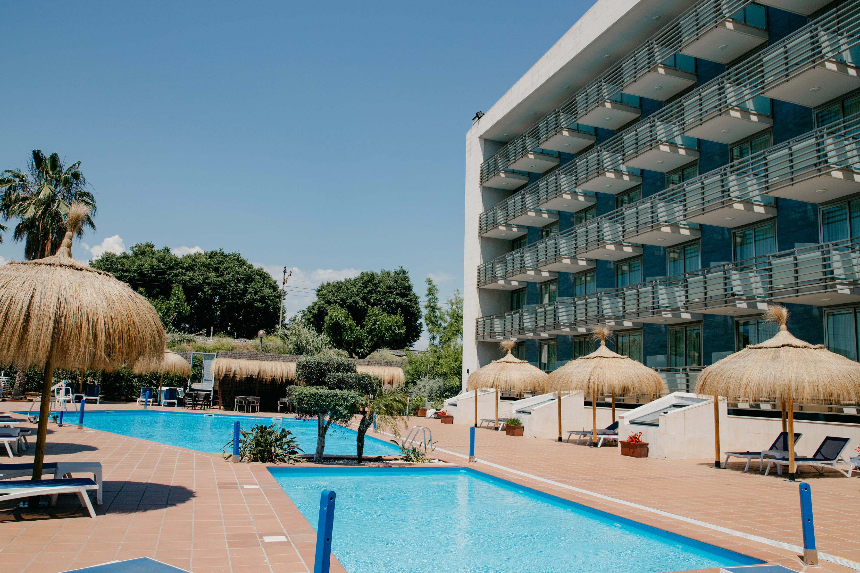 a pool with straw umbrellas and a building with trees and blue sky