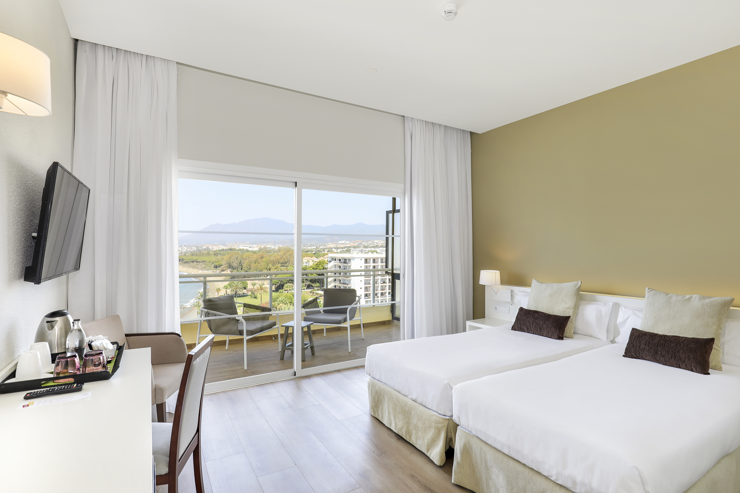 Bright hotel room with twin beds, balcony, desk, and panoramic sea view.