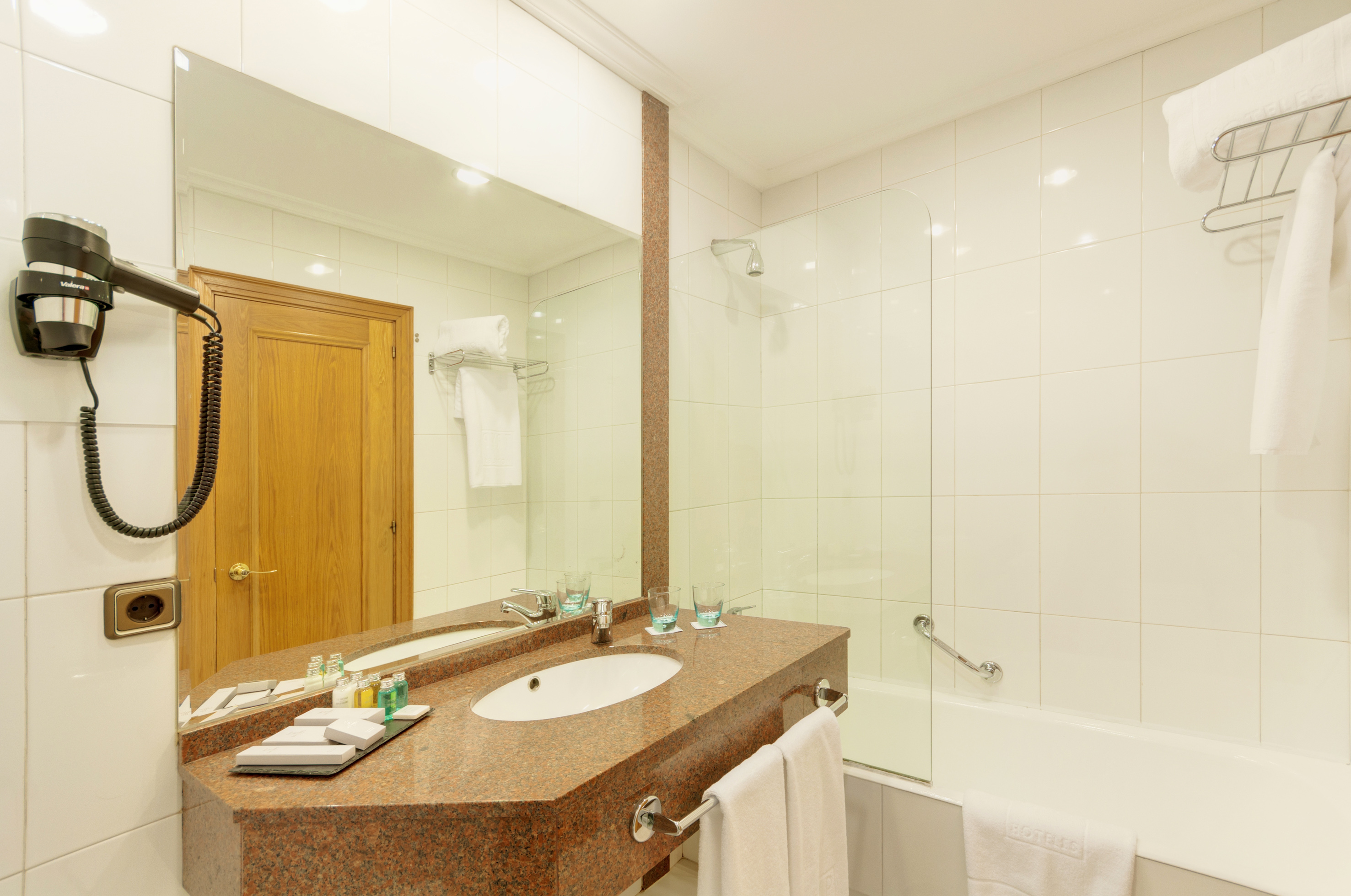 Tiled hotel bathroom with granite vanity, sink, mirror, bathtub, and hairdryer.