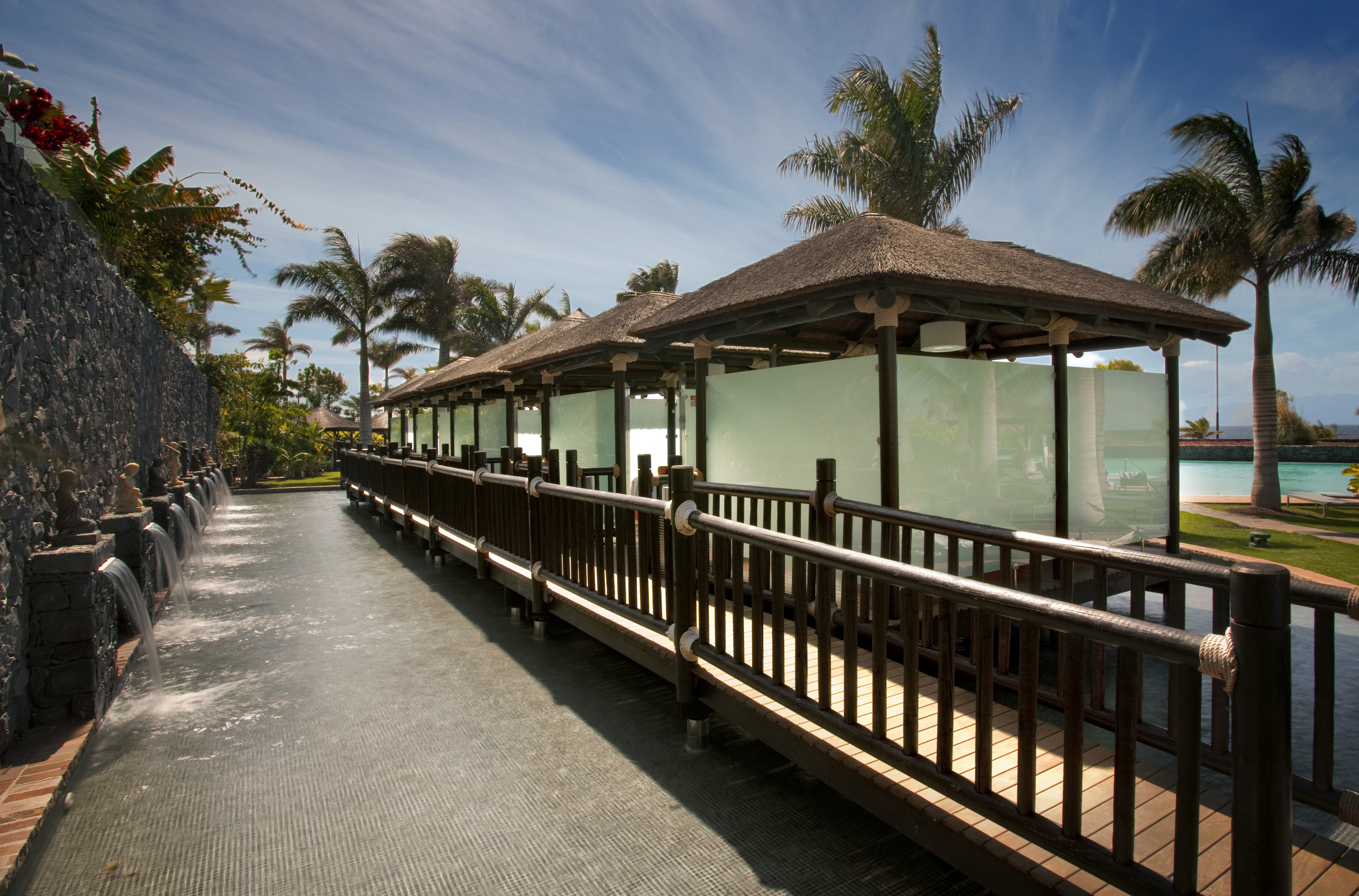 Tropical resort walkway, thatched roofs, Buddha fountains, and palm trees.