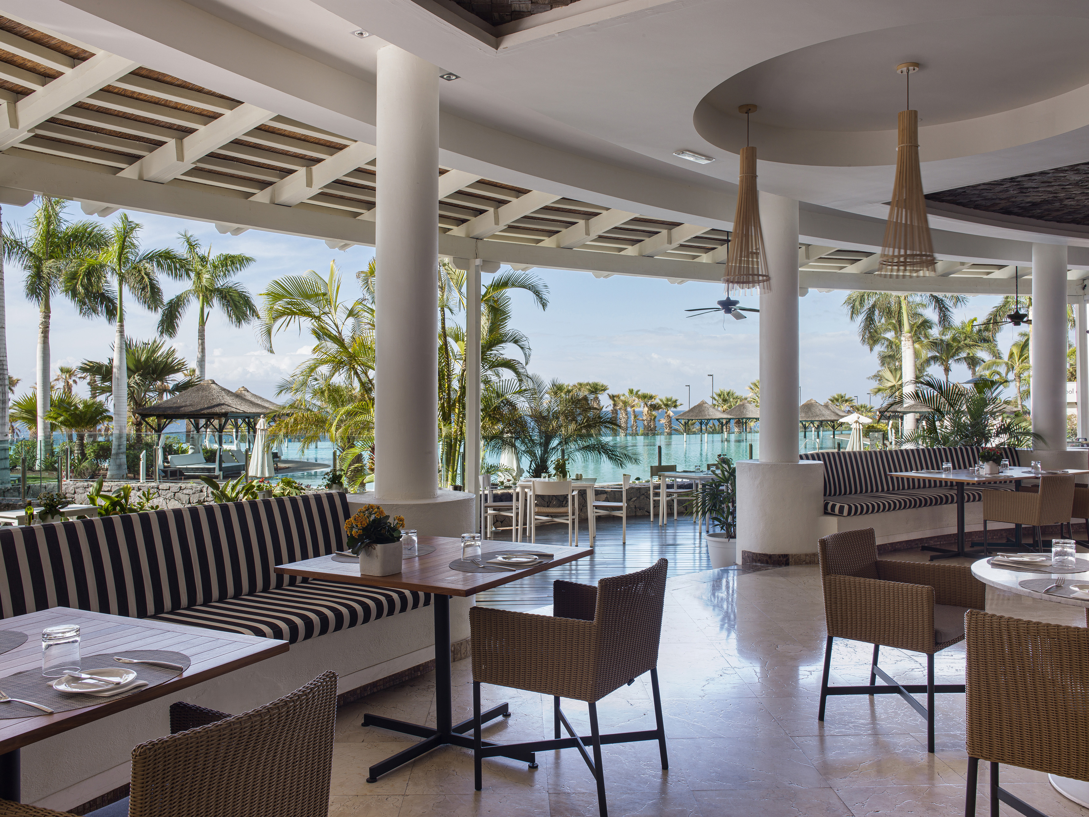 Open-air resort restaurant with pool view, striped seating, and palm trees.