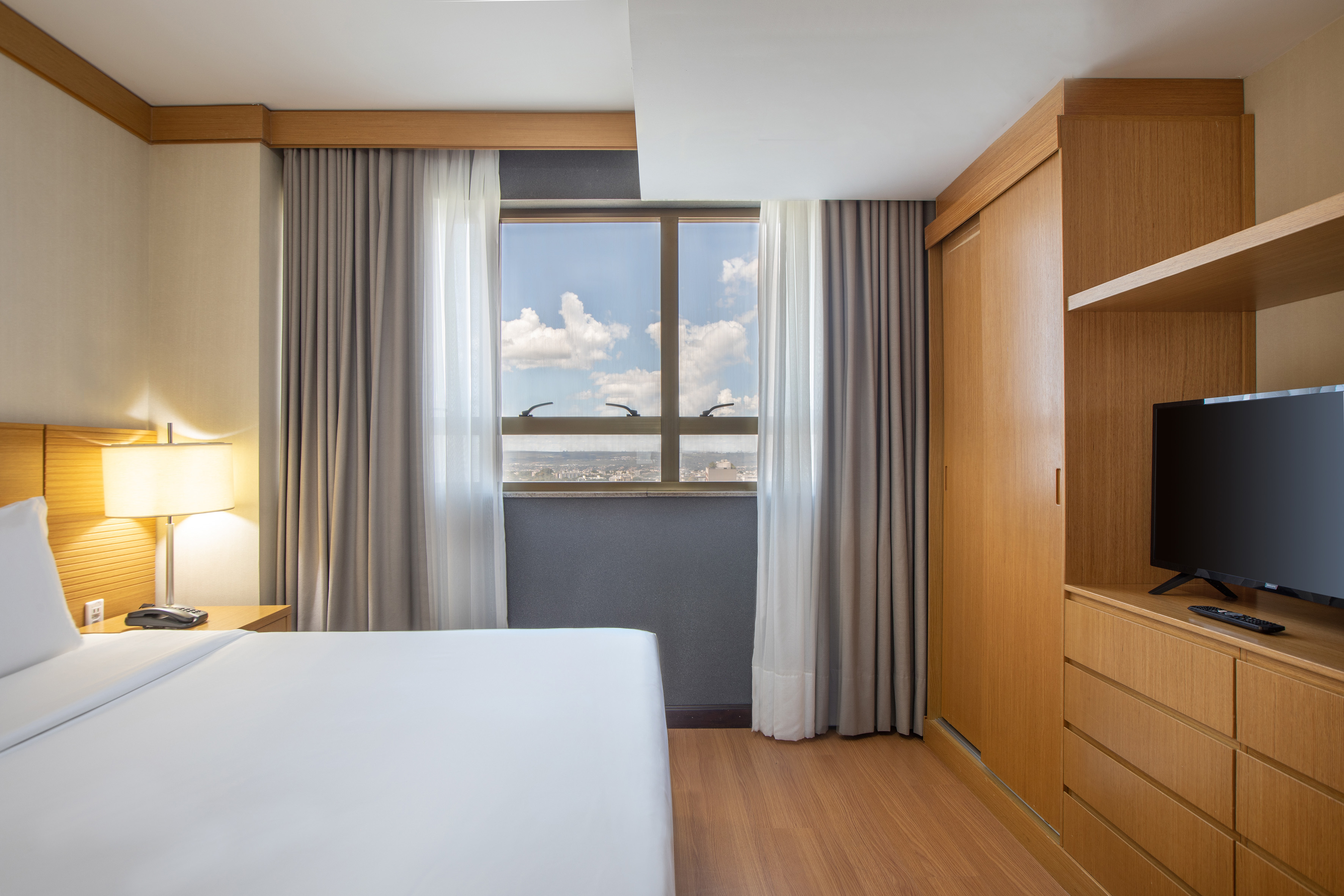 Modern hotel bedroom with white bed, city view window, wooden wardrobe, and TV.