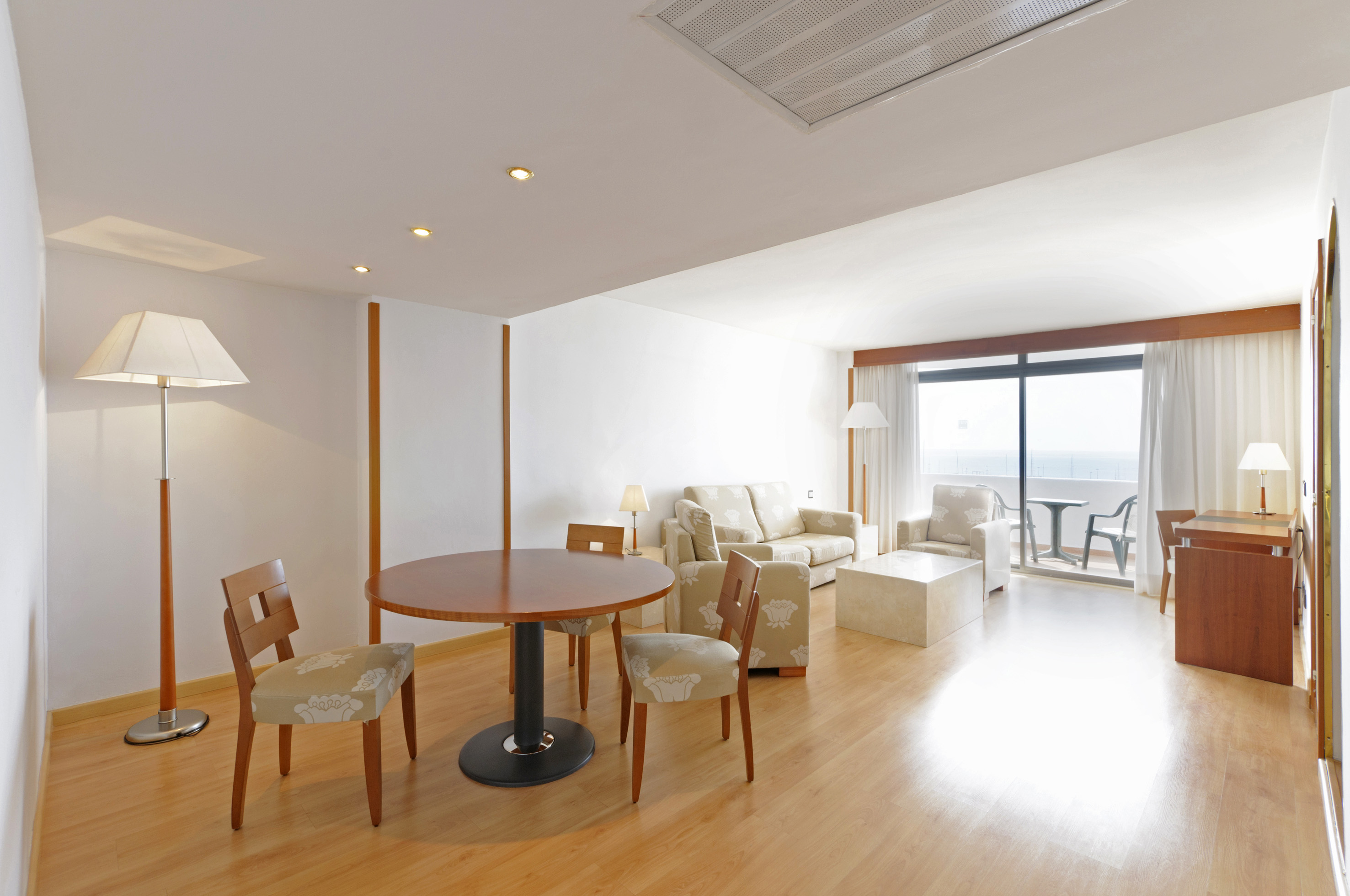 Modern hotel suite living room with sea view balcony, dining table, and light wood floor.