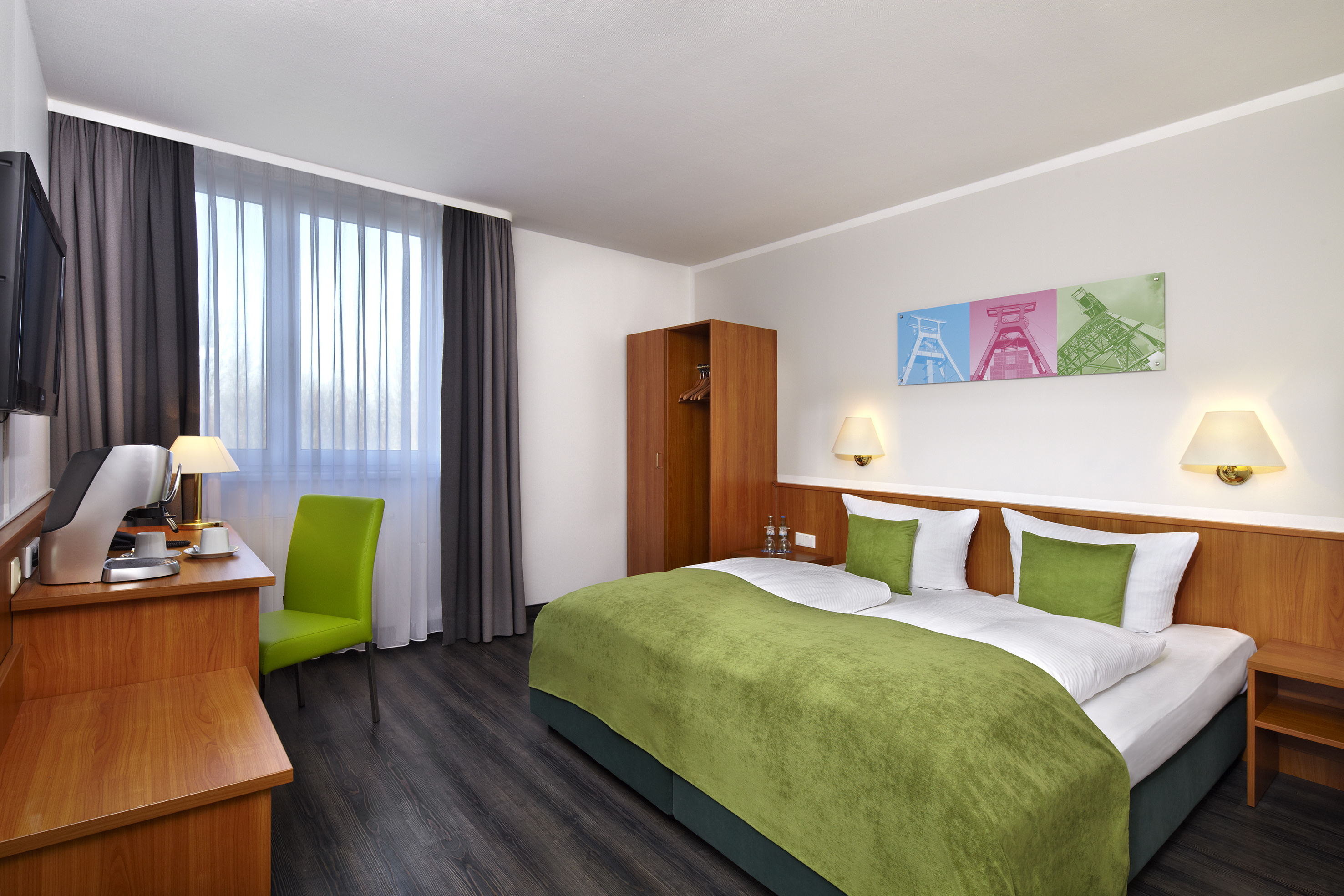 Modern hotel room with green bed, dark wood floor, desk, and industrial art.
