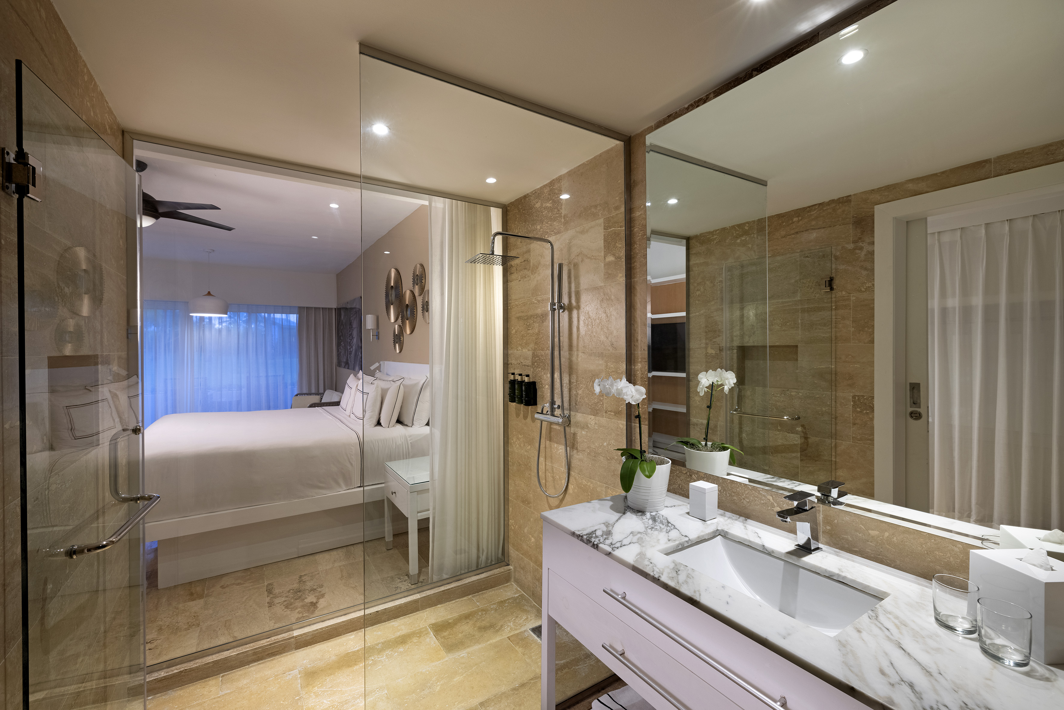 Modern hotel suite with glass shower, marble vanity, and bedroom.