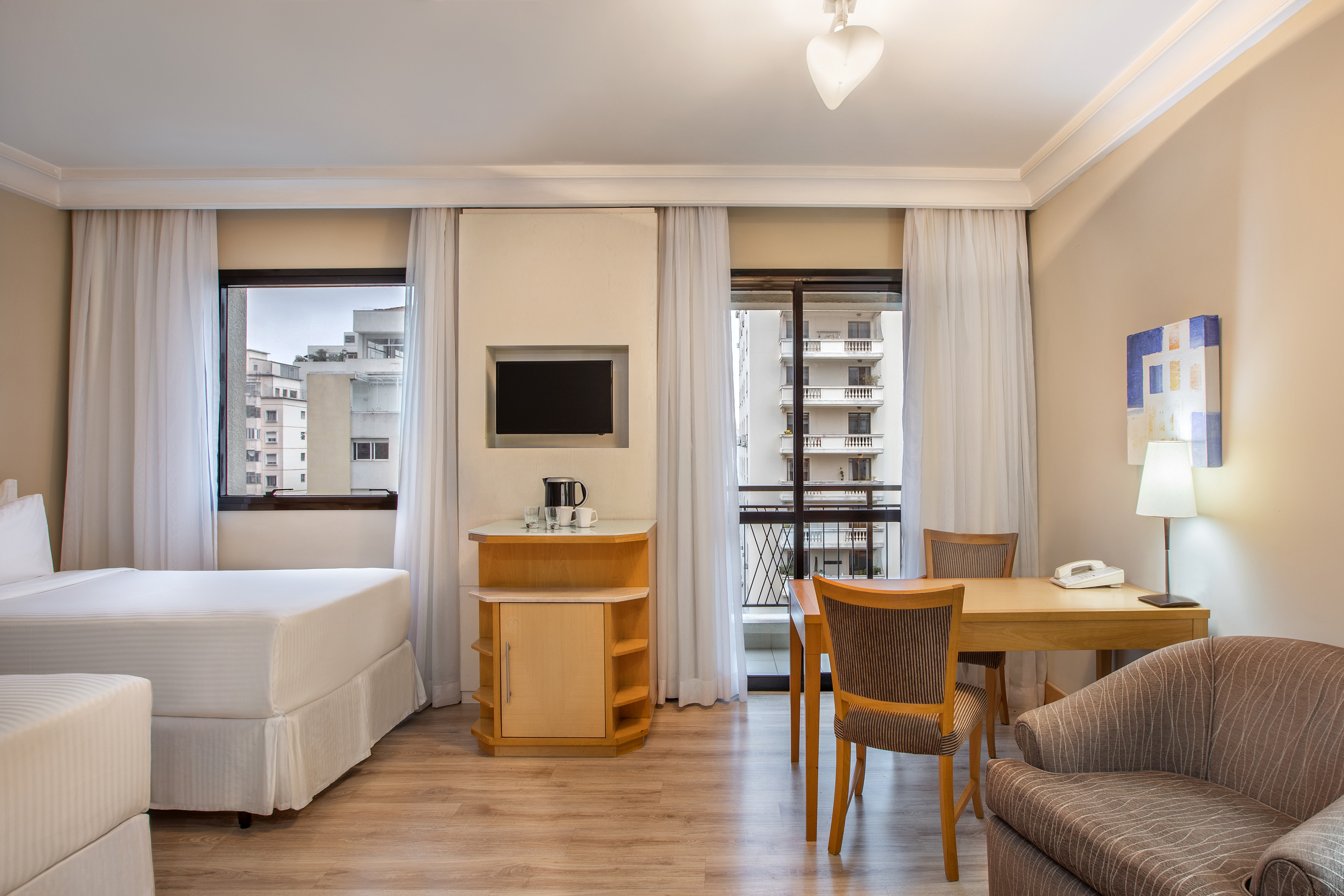 Modern hotel room with bed, dining table, TV, and city view balcony.