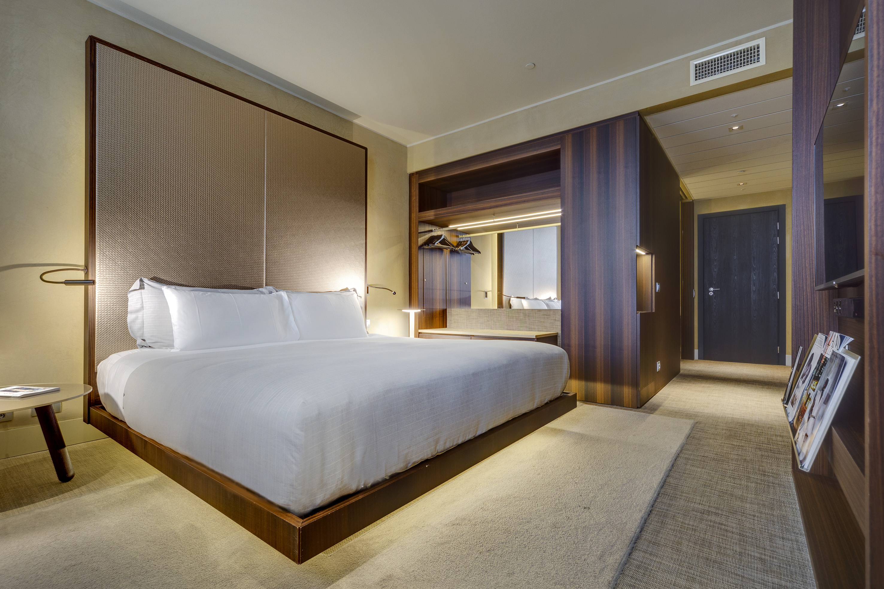 Modern hotel bedroom with king bed, textured headboard, wood paneling, and warm lighting.