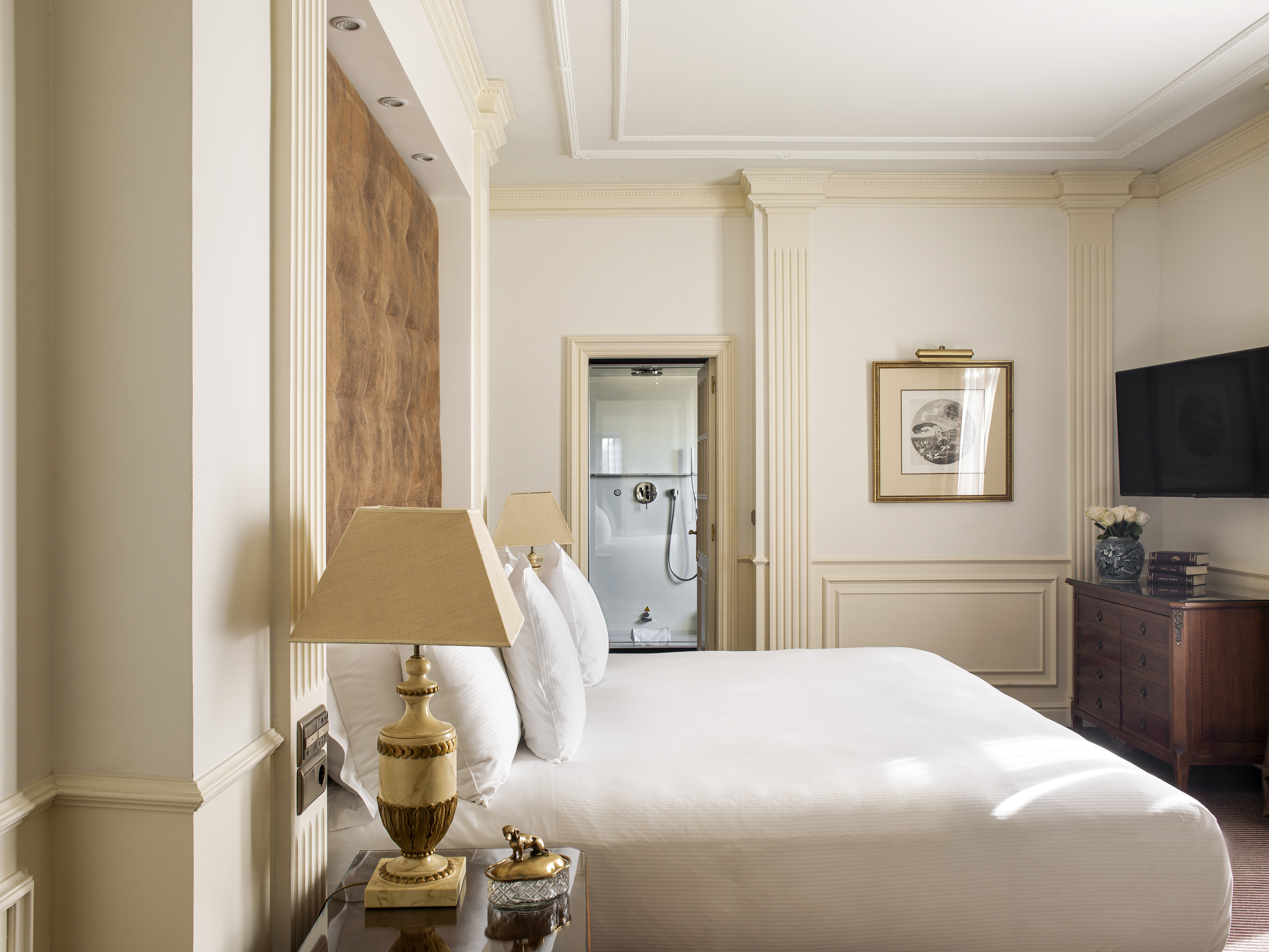 Luxurious bedroom with white bed, ornate moldings, and glass shower.