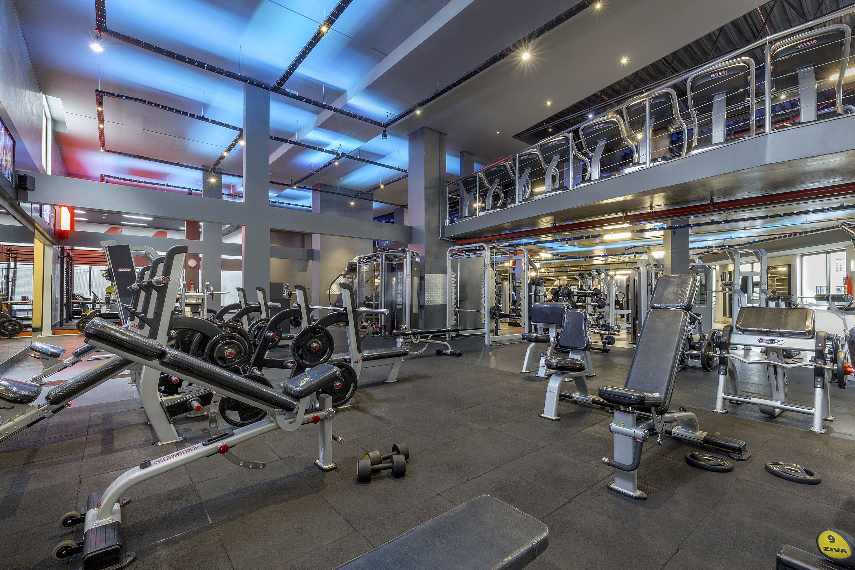 Modern gym with fitness equipment, blue lighting, and mezzanine level.