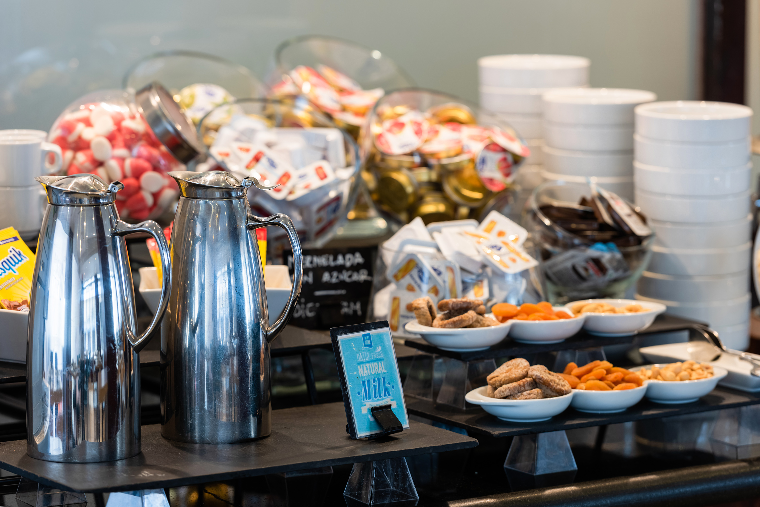 Breakfast buffet display with thermoses, dried fruits, nuts, cookies, and milk sign.