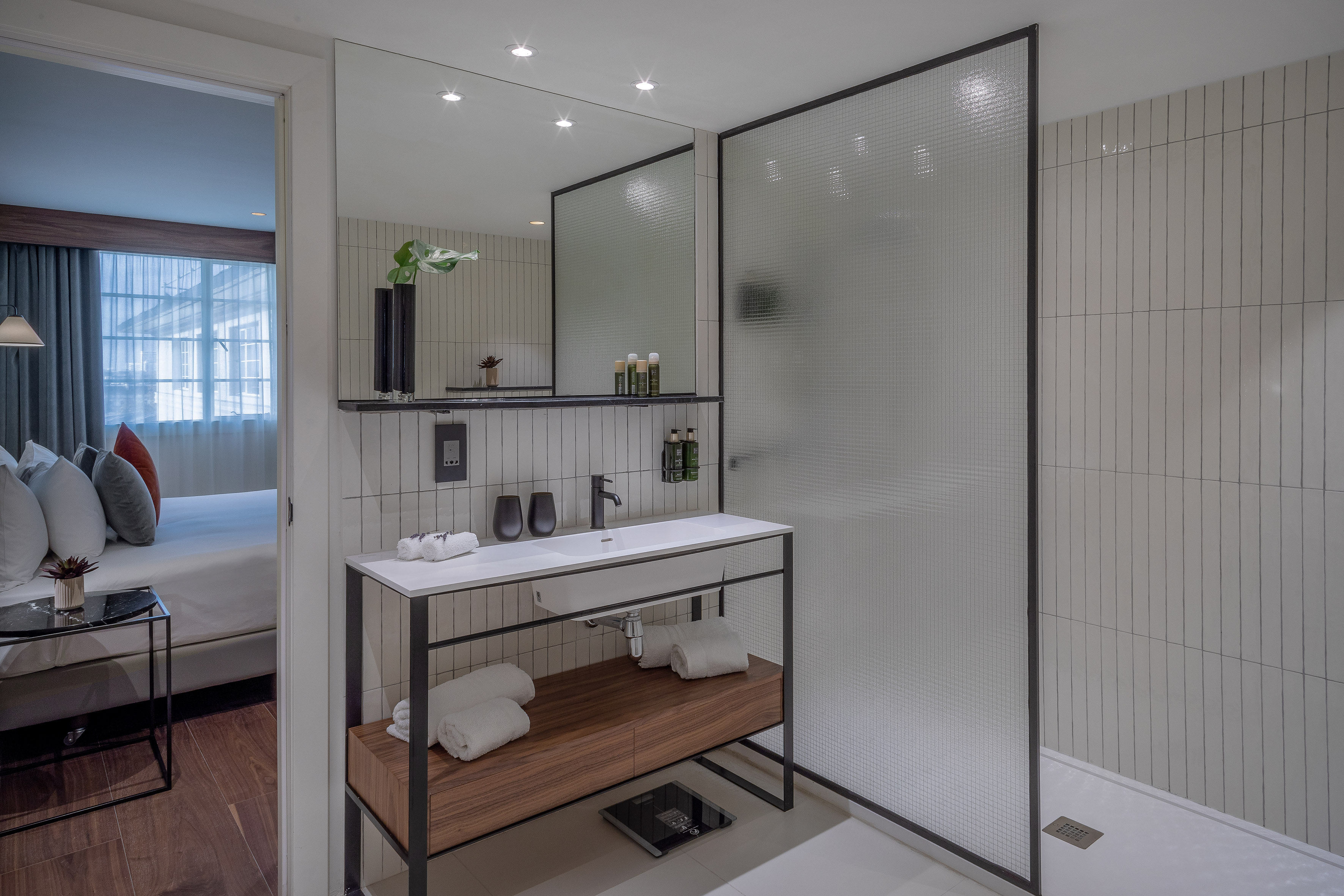Modern hotel bathroom with vanity, mirror, glass shower, and connecting bedroom.