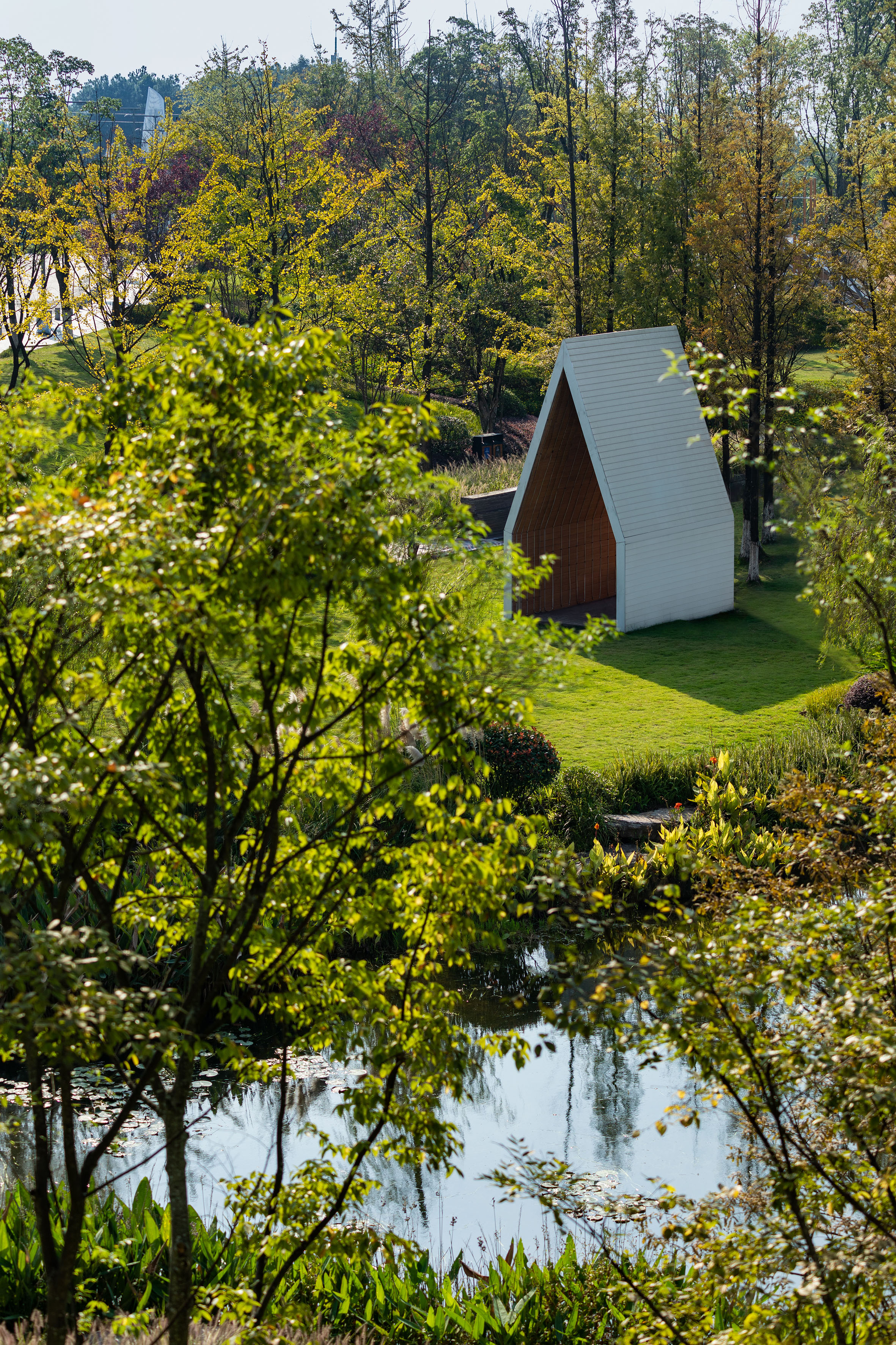 Modern A-frame cabin beside a pond, surrounded by green trees and lawn.
