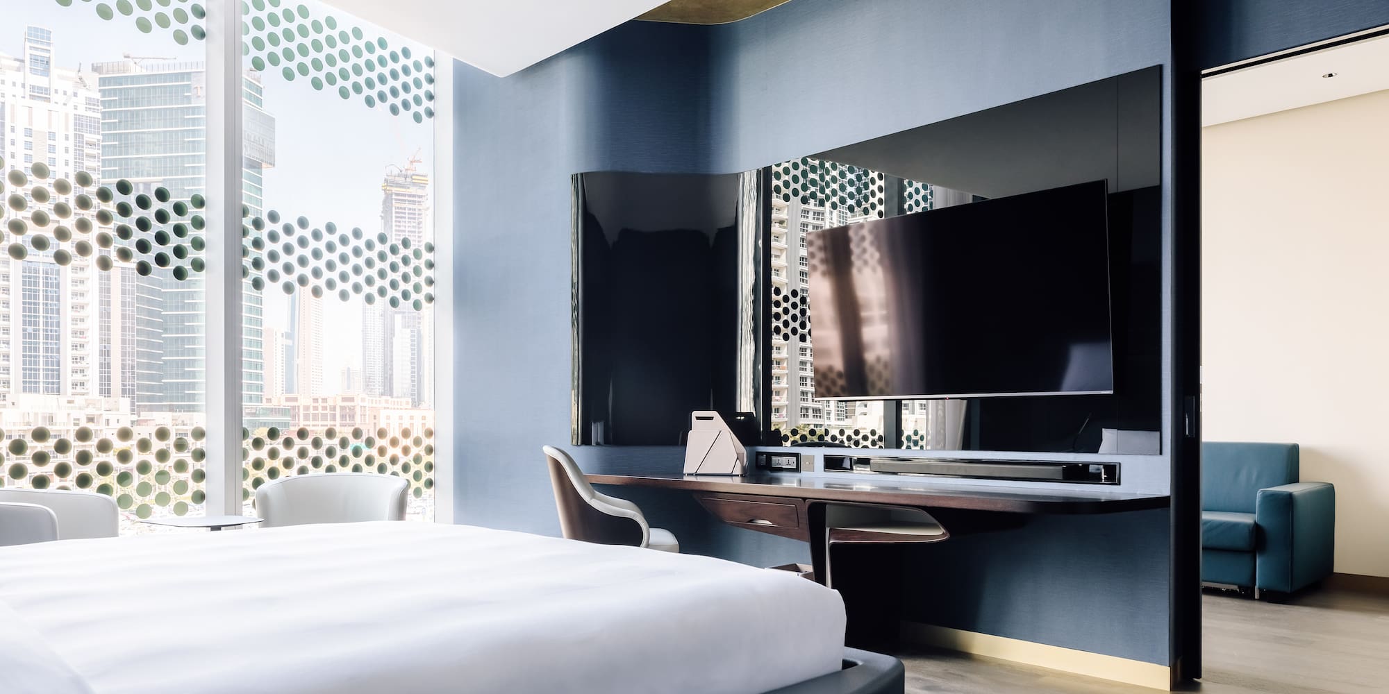 Modern hotel suite bedroom with city view, patterned windows, large bed, and TV.