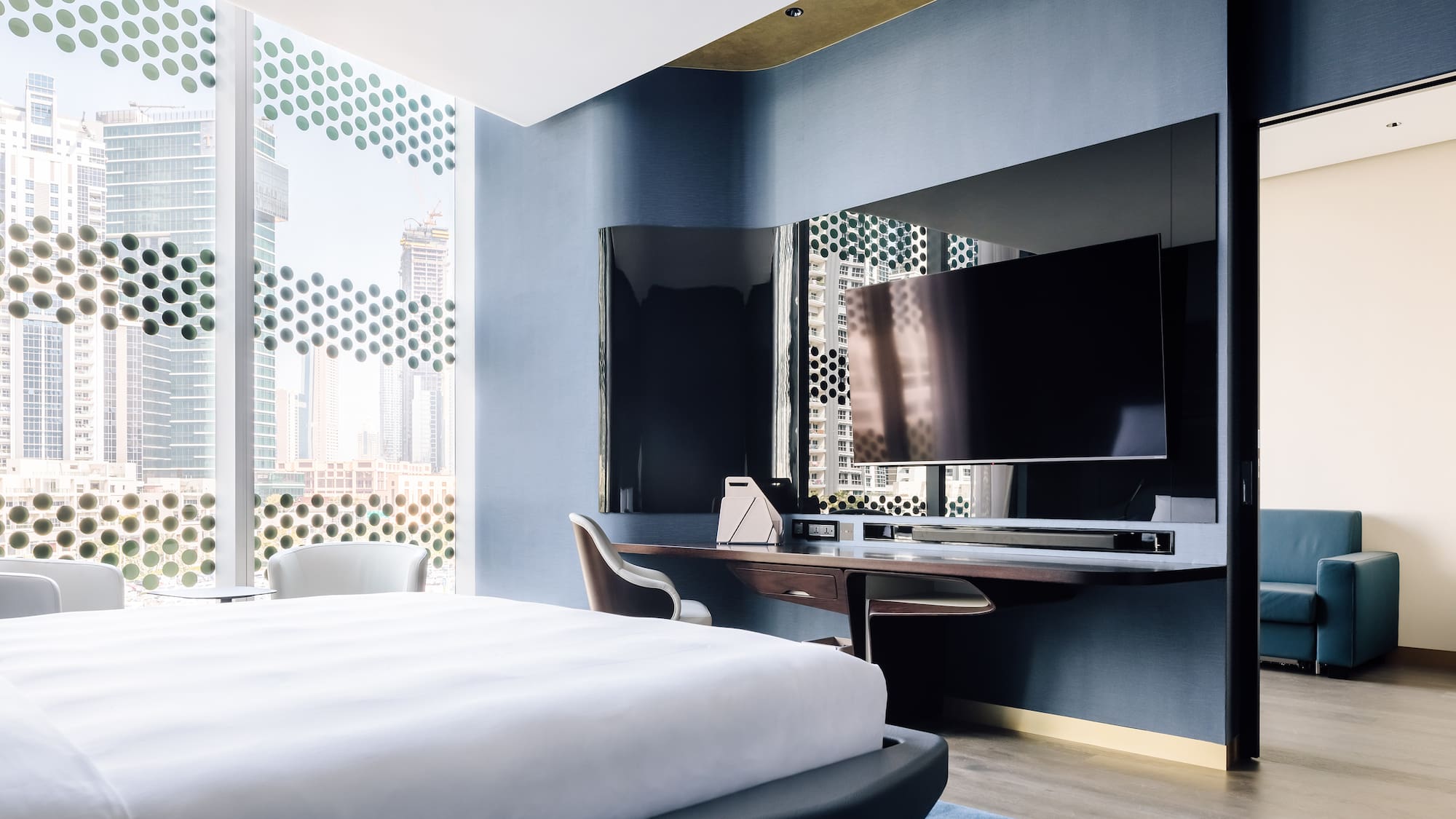 Modern hotel suite bedroom with city view, patterned windows, large bed, and TV.