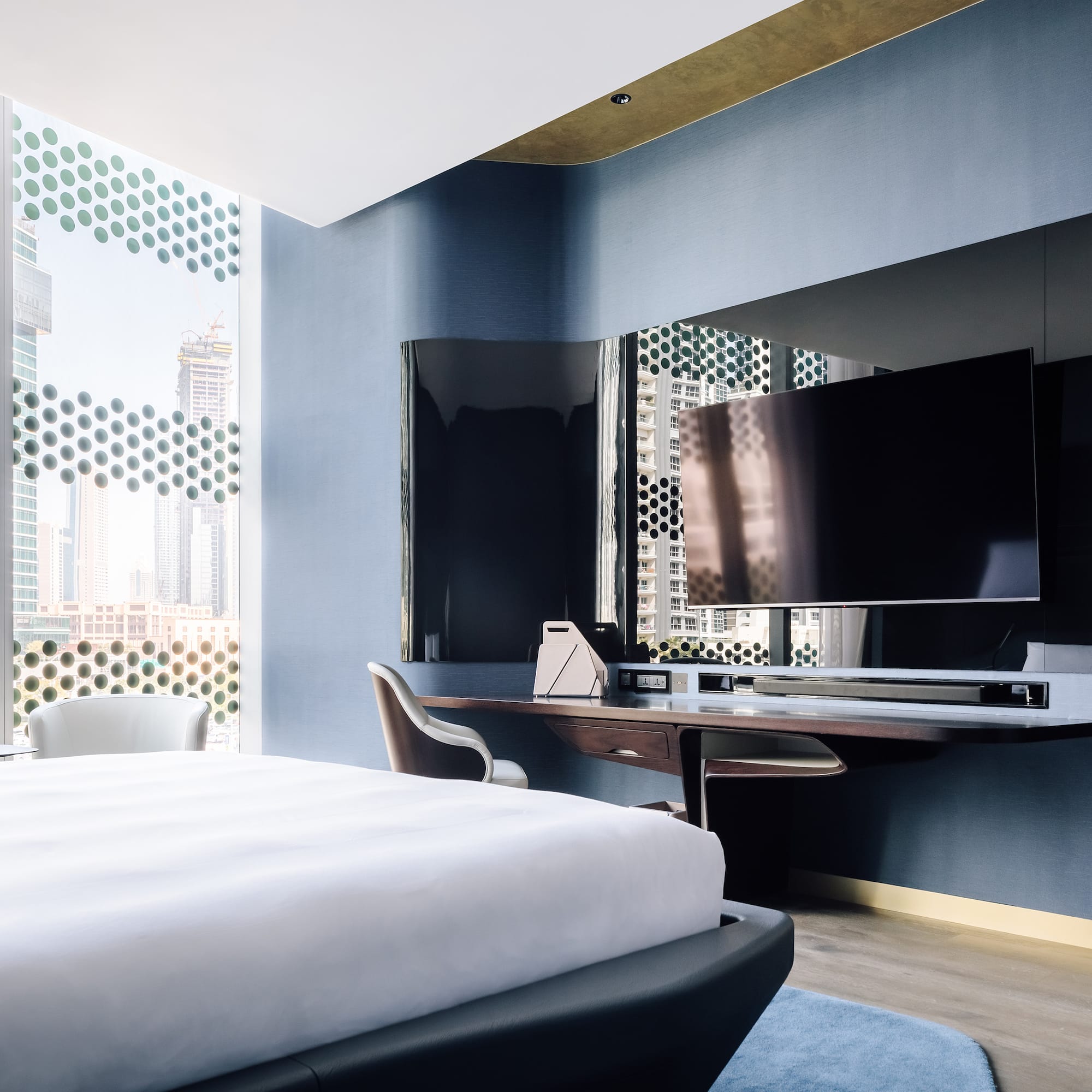 Modern hotel suite bedroom with city view, patterned windows, large bed, and TV.
