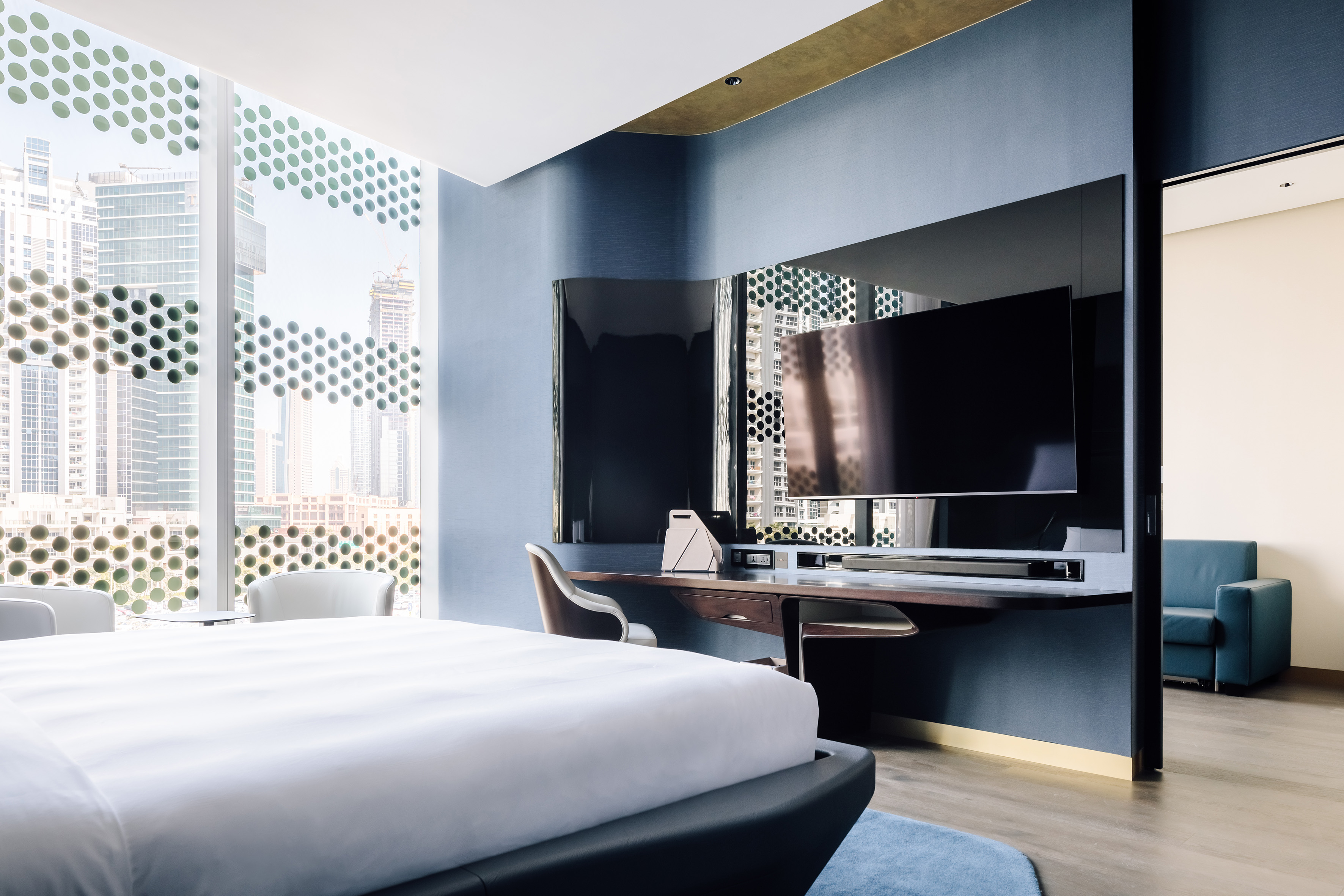 Modern hotel suite bedroom with city view, patterned windows, large bed, and TV.