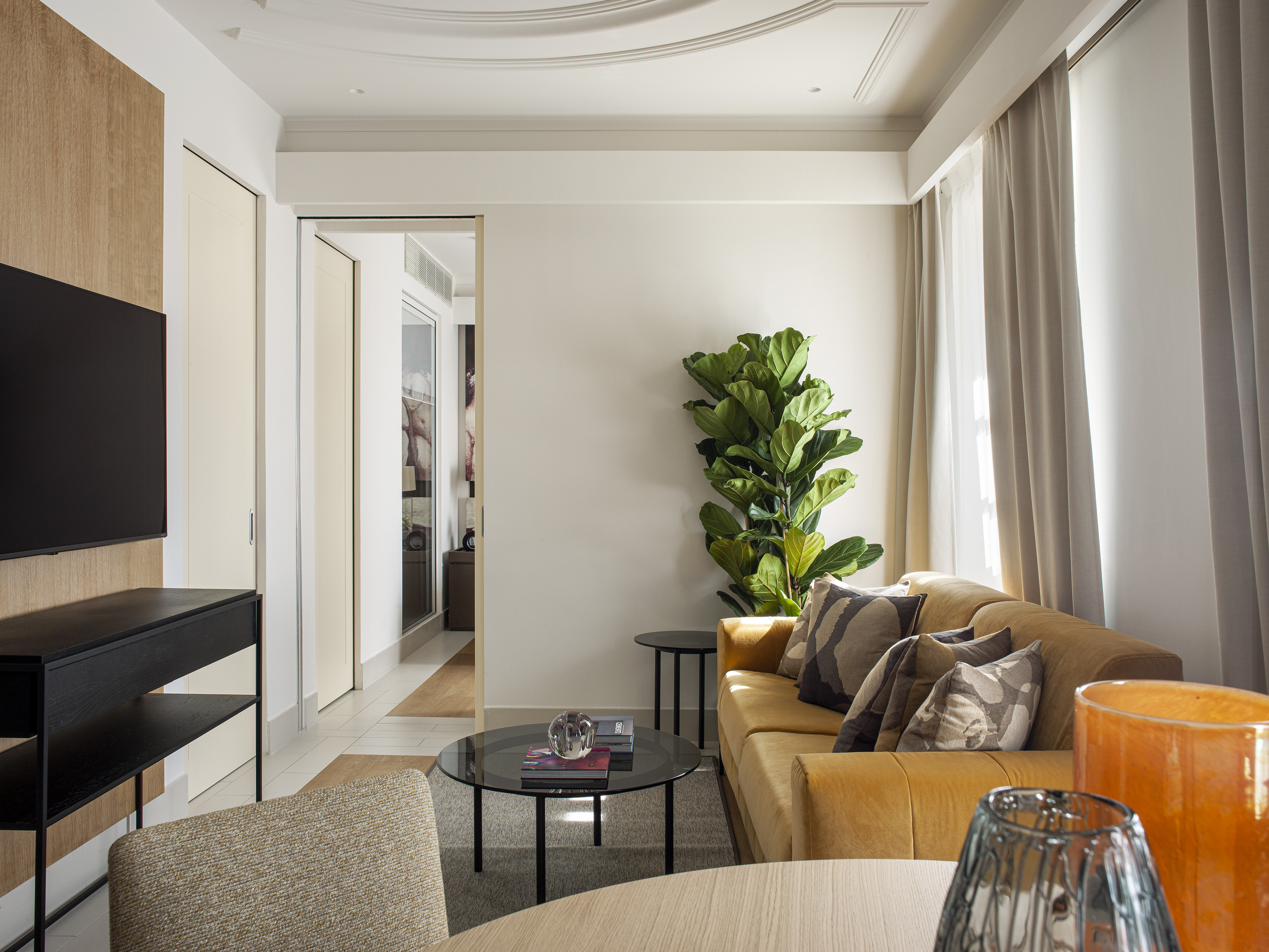 Elegant living room with golden sofa, glass coffee table, large fiddle leaf fig, and TV.
