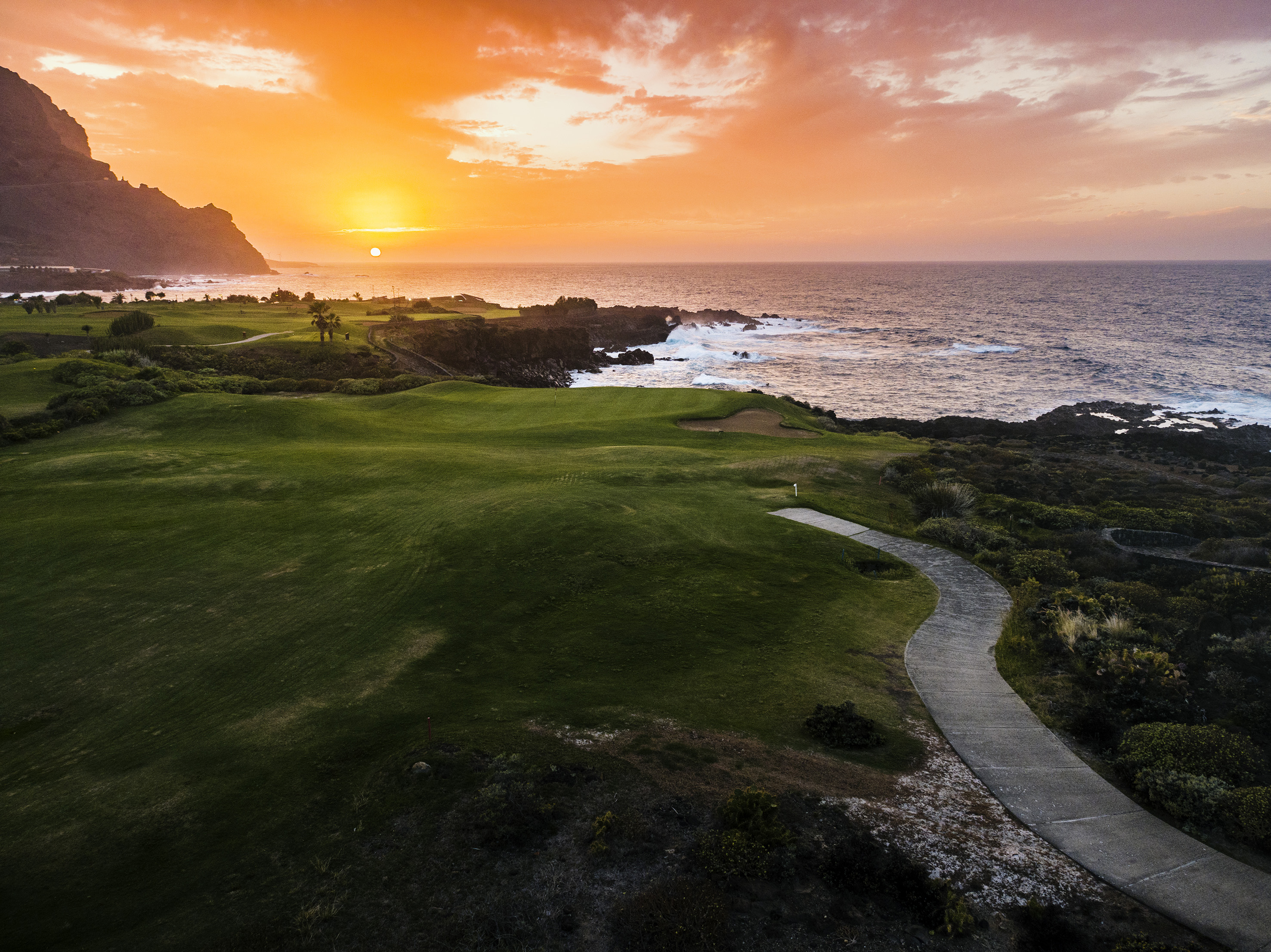 Oceanfront golf course sunset, vibrant orange sky, green fairways, rocky coast.