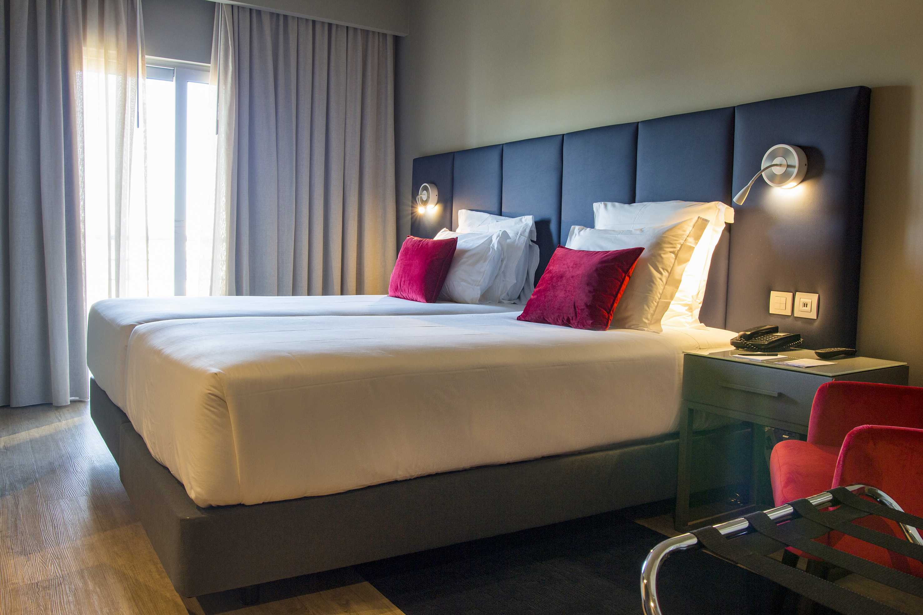 Modern hotel room with twin beds, blue headboard, red pillows, and warm lighting.