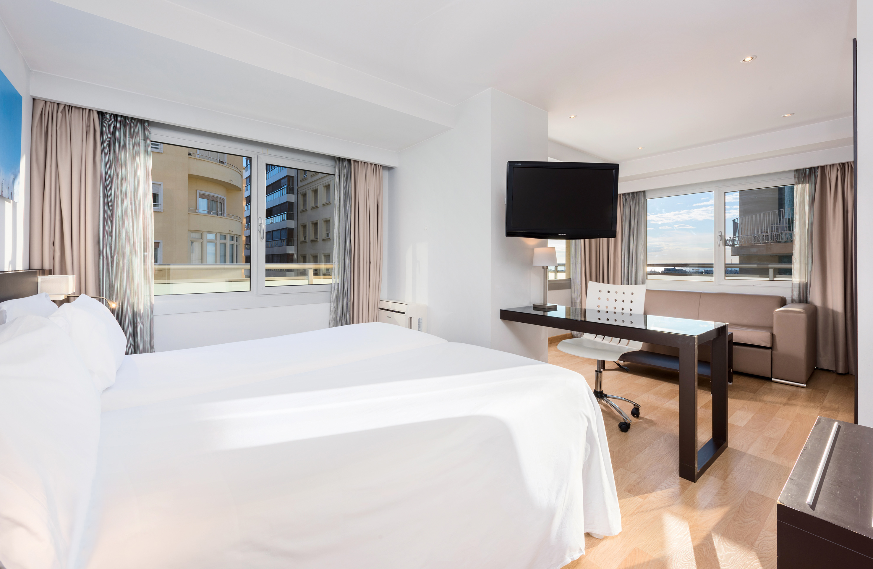 Modern hotel room with two beds, desk, TV, sofa, and city view.