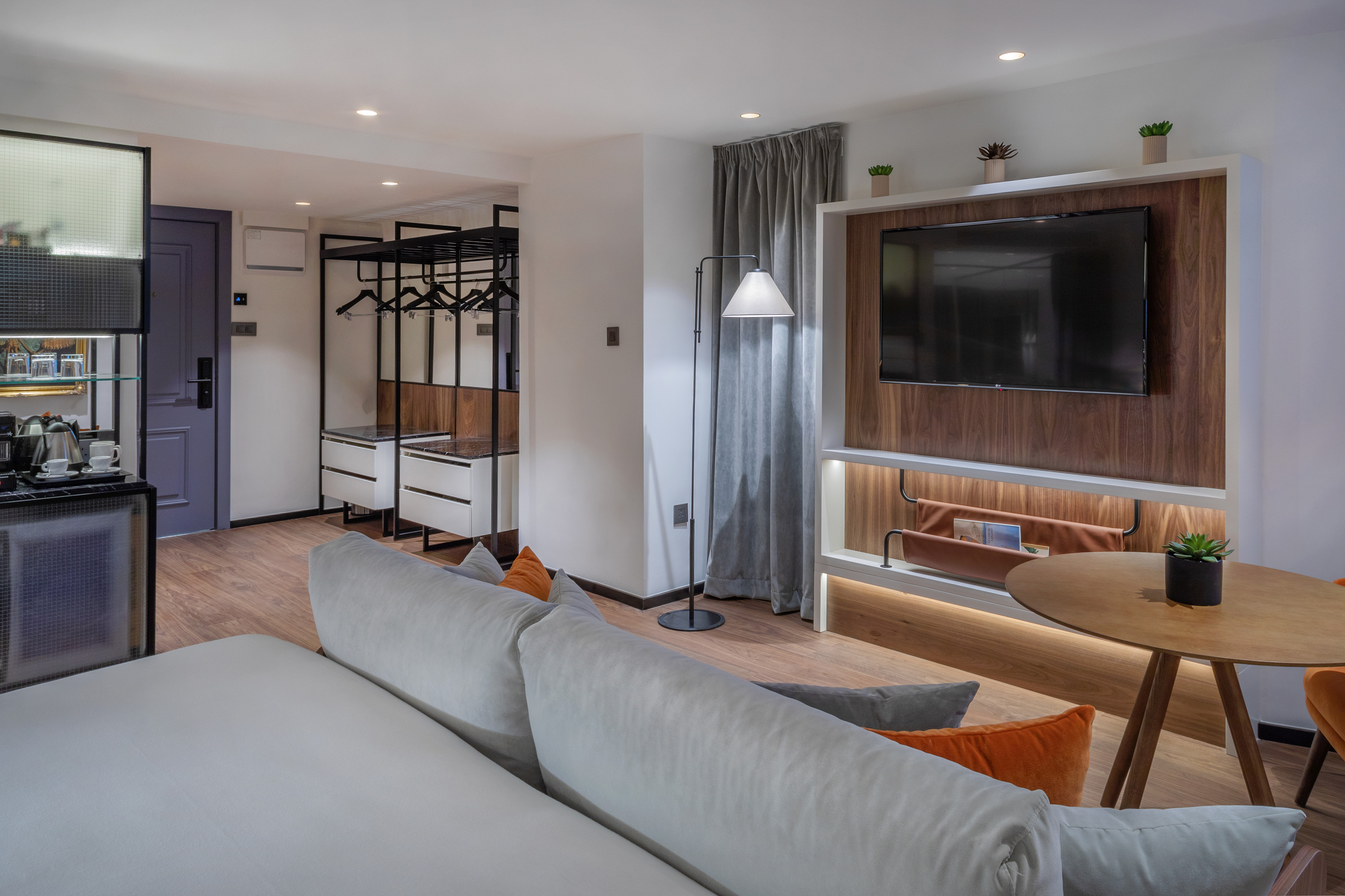 Modern hotel suite with living space, TV, kitchenette, and open wardrobe.