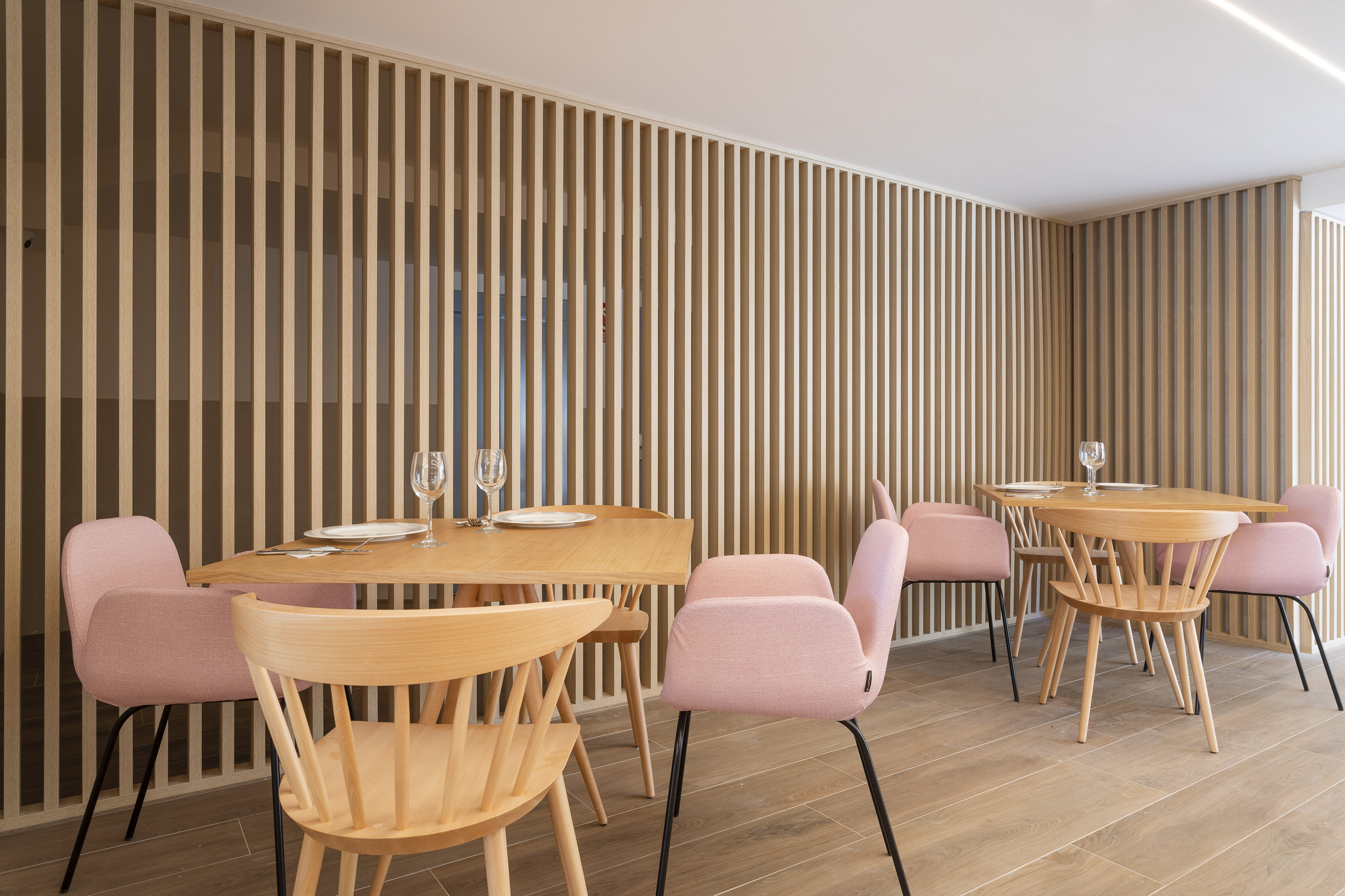 Modern restaurant dining with wood slat walls, set tables, and pink chairs.