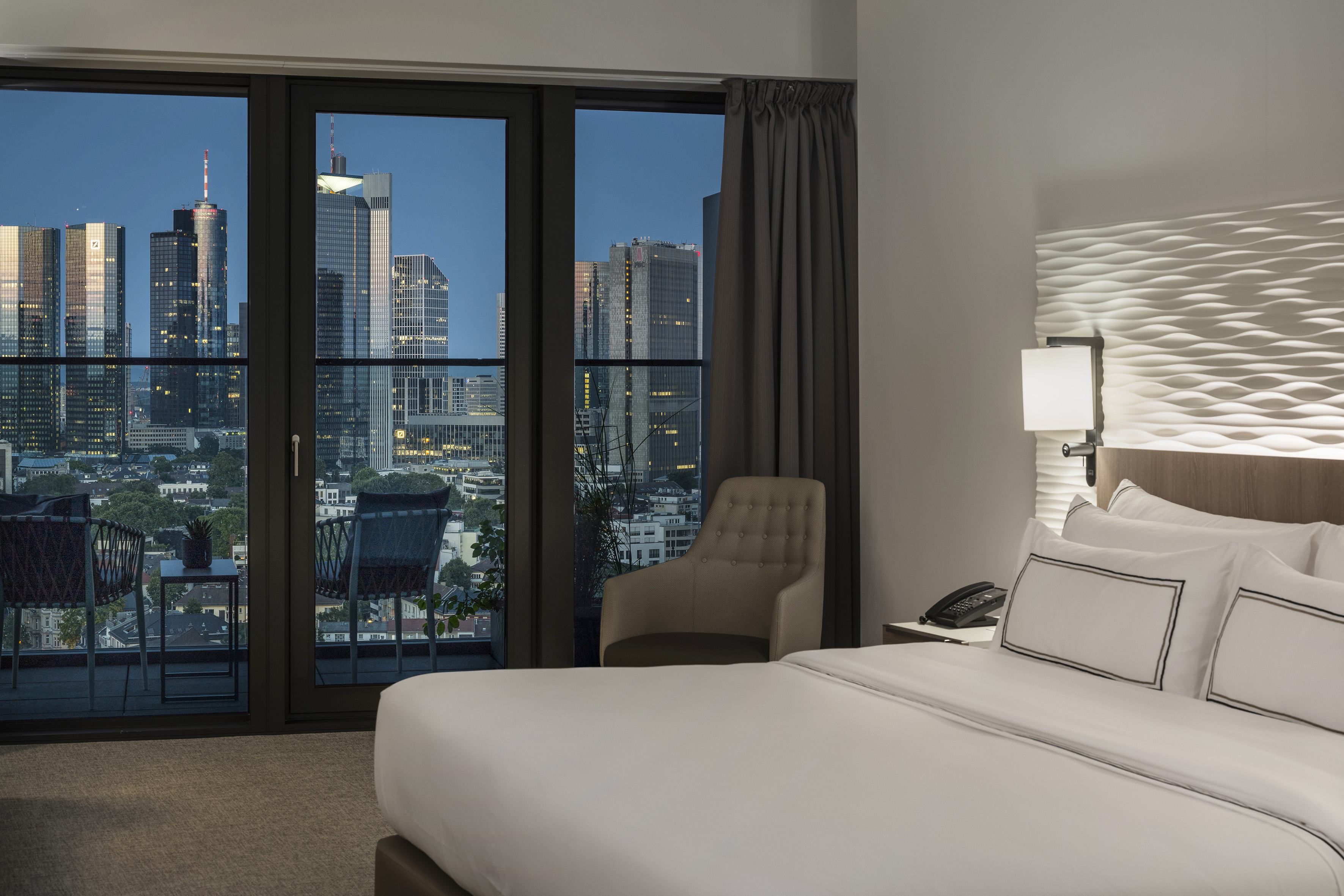 Modern hotel room with evening city skyline view, balcony, and comfortable bed.