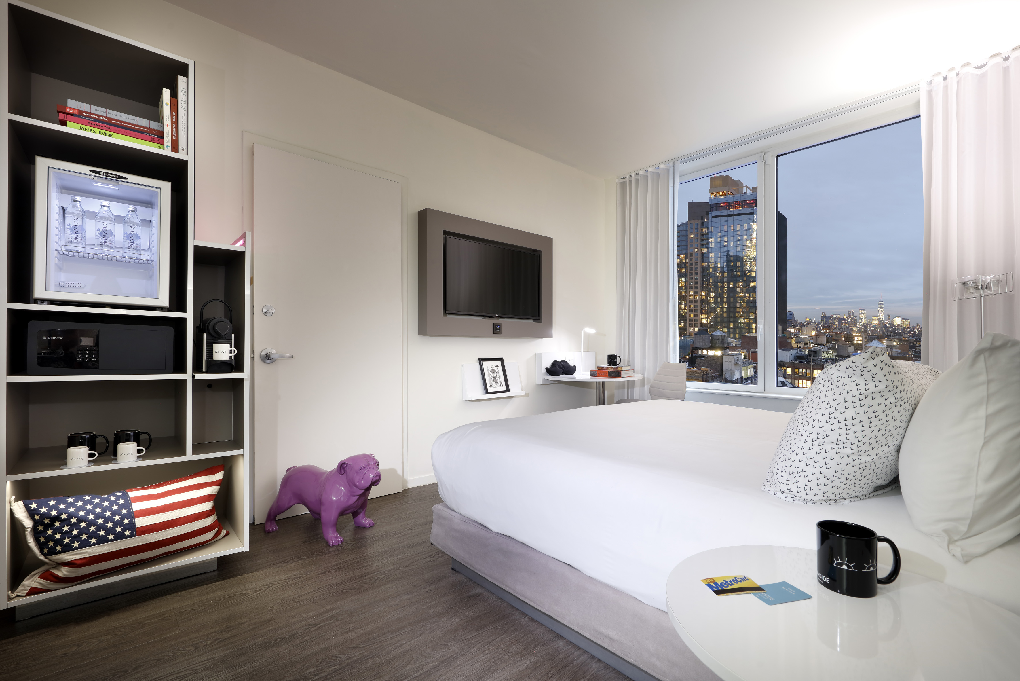 Modern hotel room with NYC skyline view, large bed, TV, and contemporary decor.