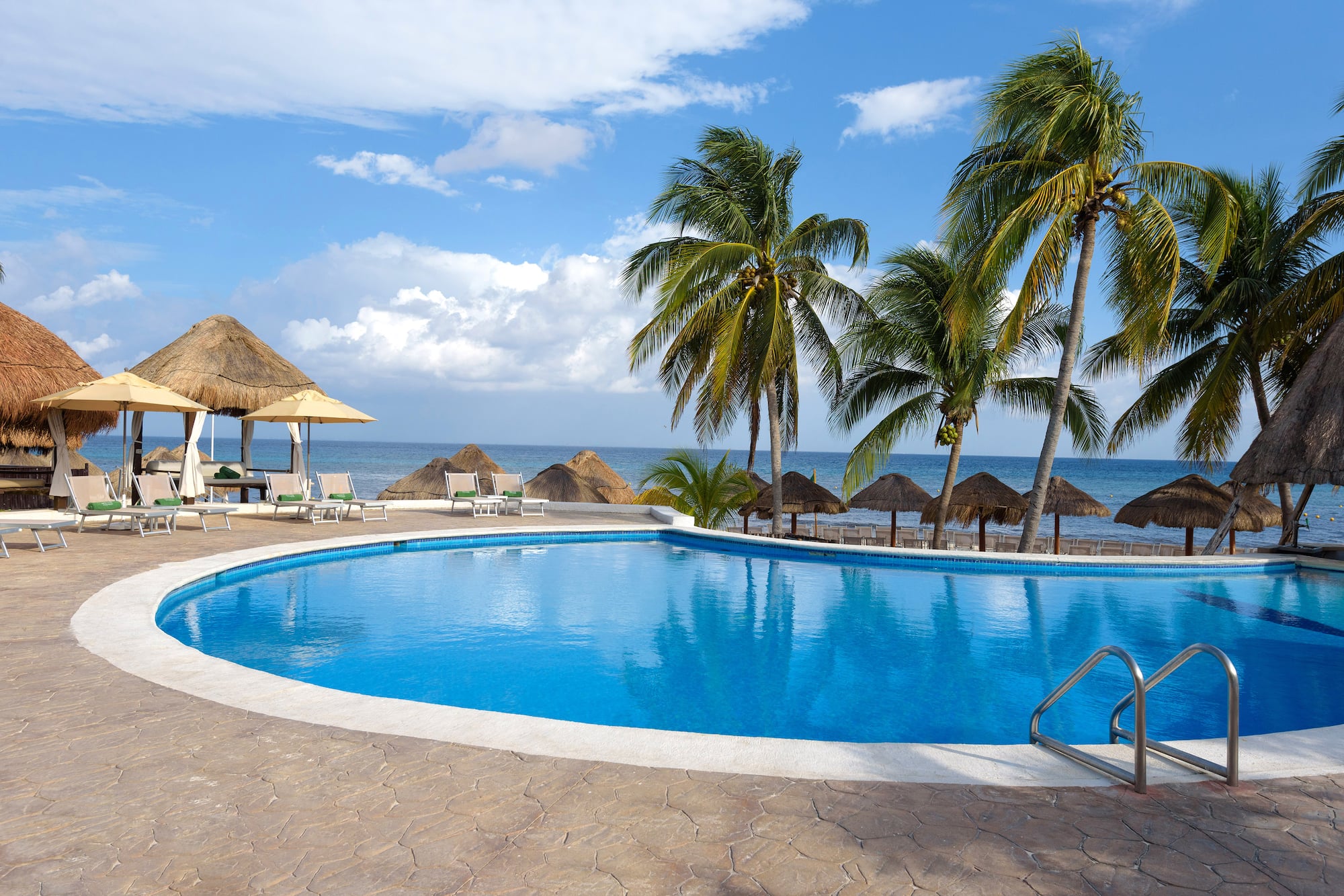 Piscina de resort tropical con palmeras, mar y cabañas de playa.