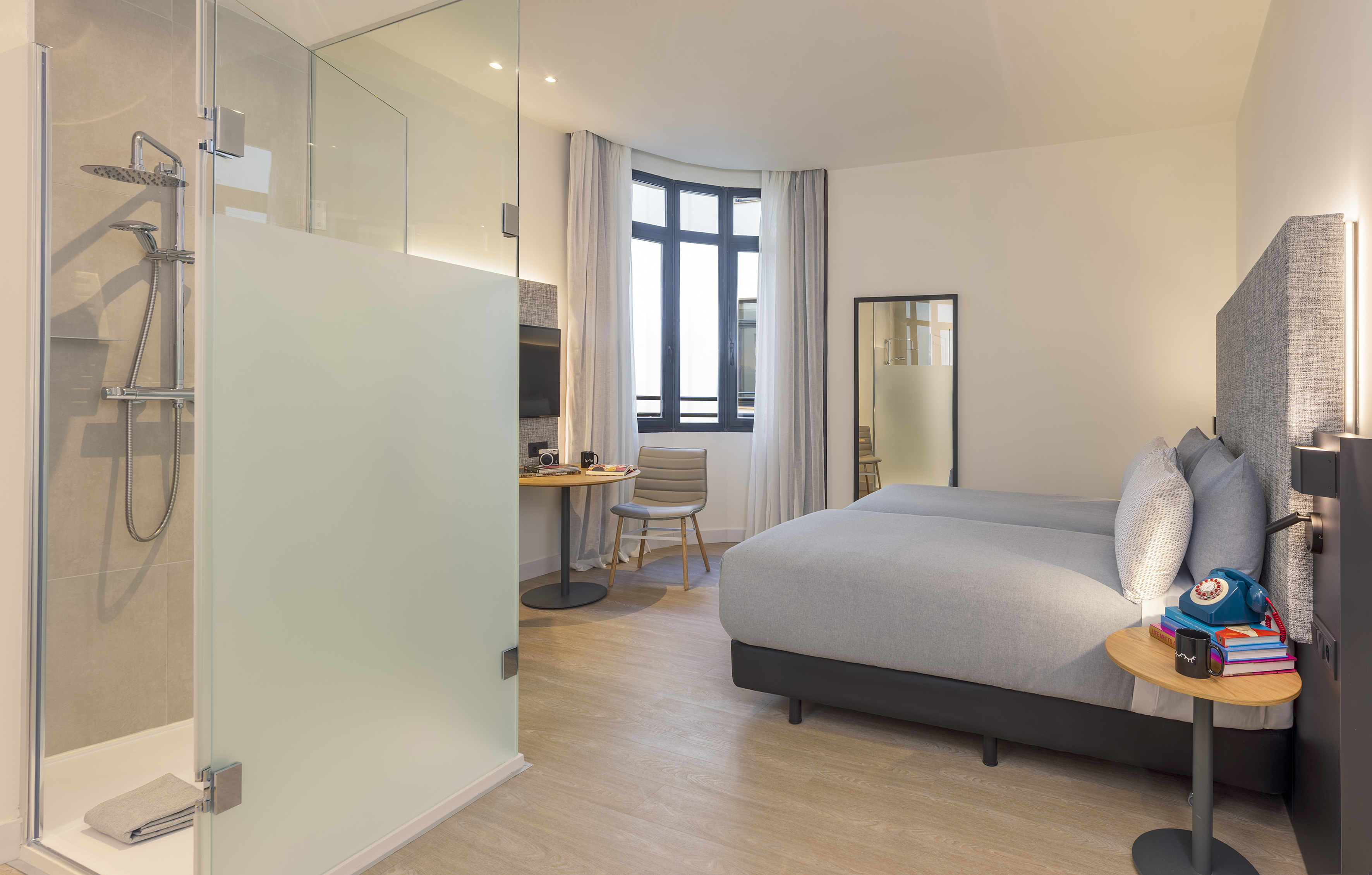 Modern hotel room with twin beds, glass shower, desk, and large window.