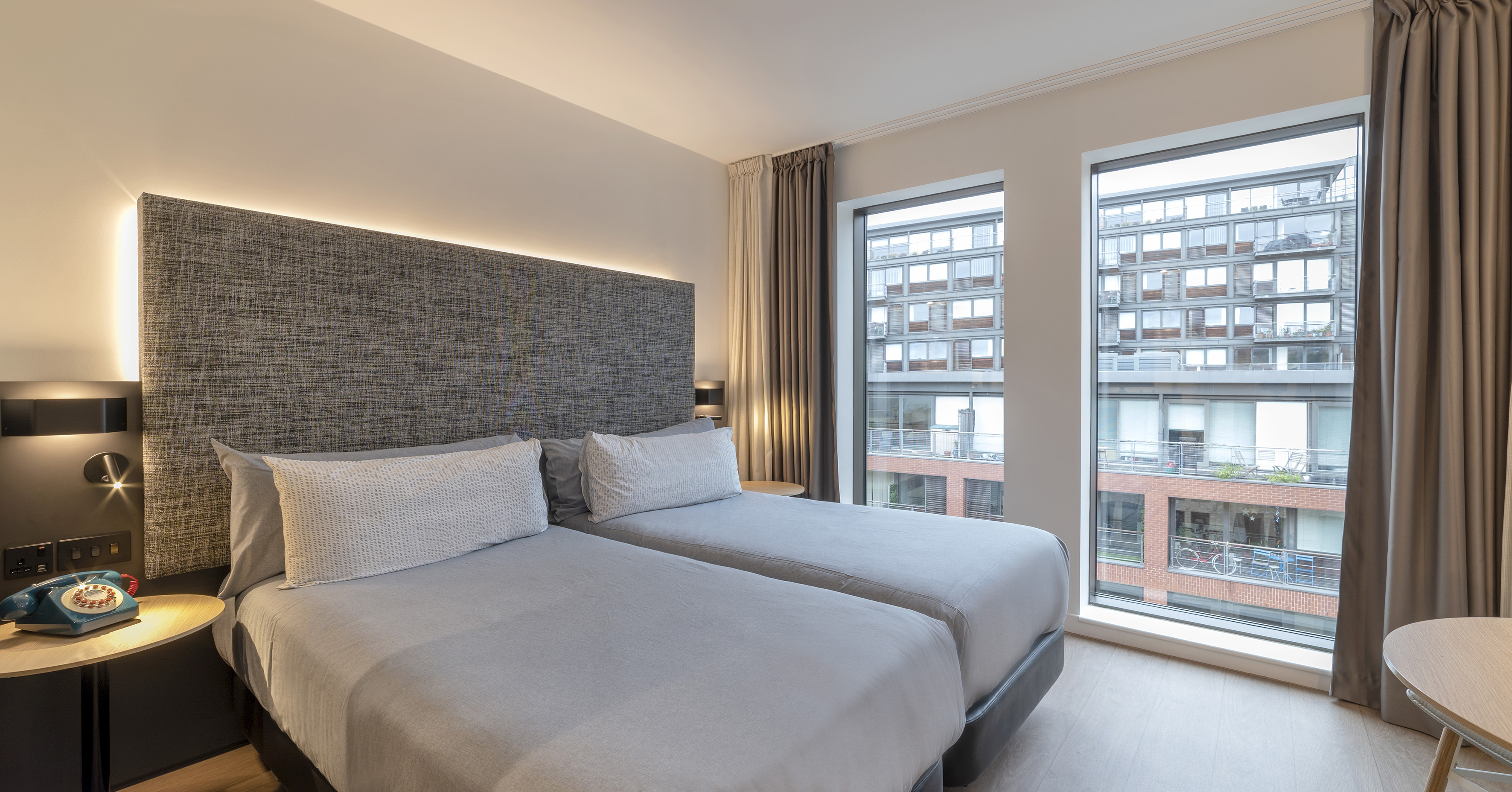 Modern twin hotel room with grey beds, backlit headboard, and city view.
